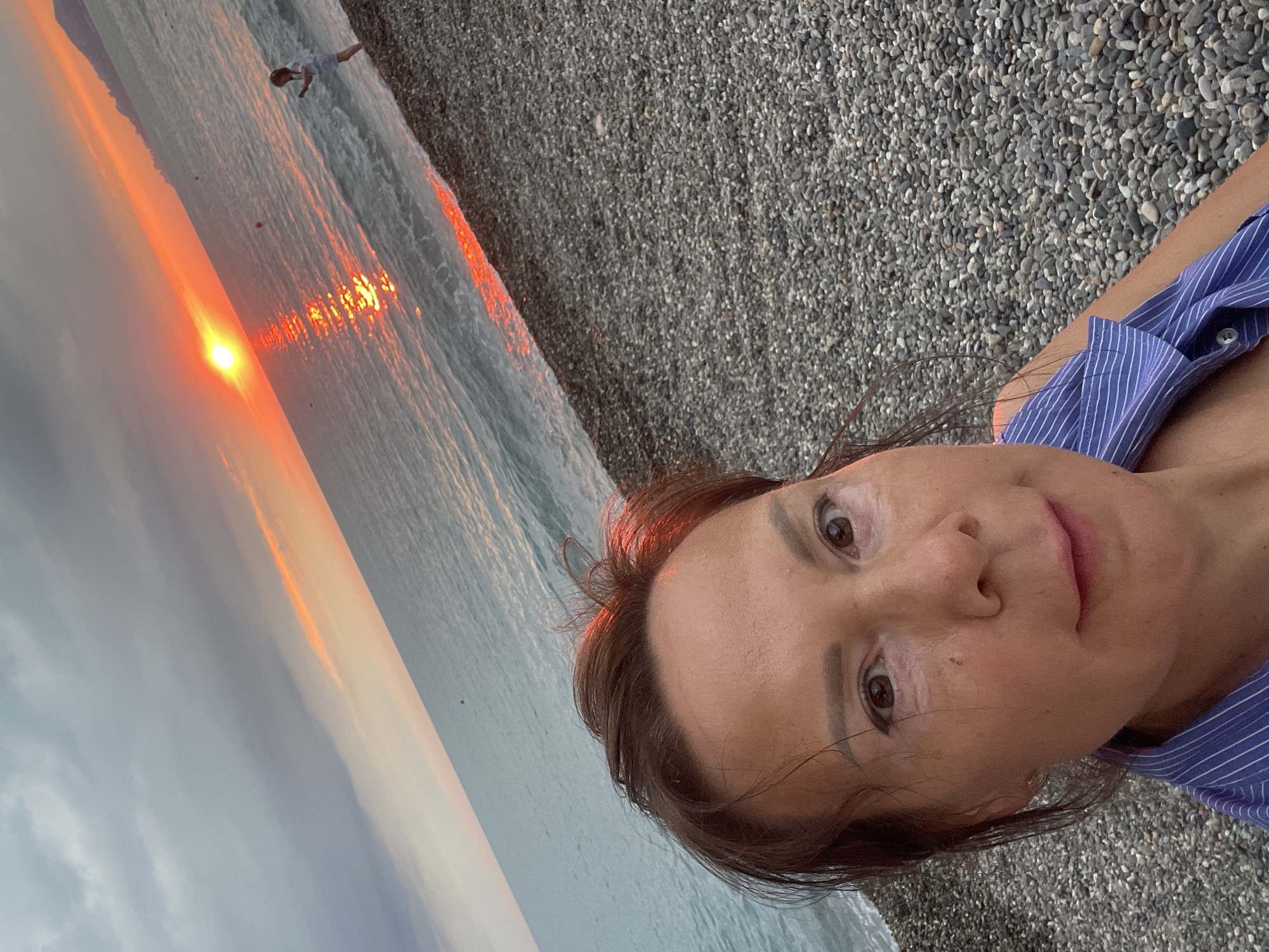 A woman is taking a selfie on a pebble beach in Sochi, Russia, with a sunset over the Black Sea in the background. The sun is low on the horizon, casting a bright orange and yellow glow on the water. A young child is visible in the distance, playing near the water's edge. The sky is cloudy with streaks of pink and orange. The woman is wearing a blue and white striped shirt.