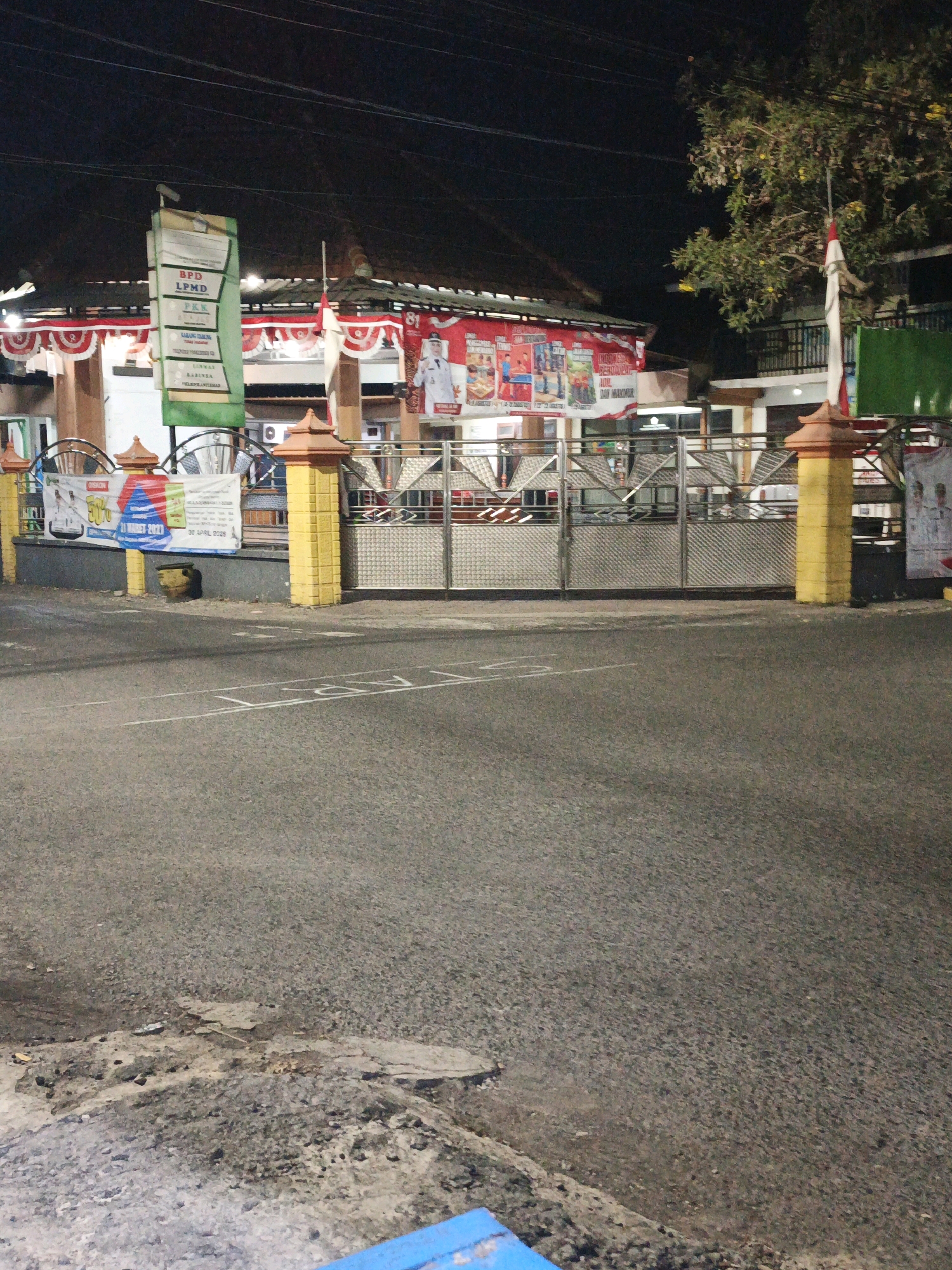 The image depicts a street scene at night in Ketegan, Indonesia. In the foreground, a paved road shows some wear and tear, with a raised, rocky embankment on the left. Across the road, there is a fenced entrance to a building, with decorative yellow pillars topped with reddish-brown accents. A large metal gate with an intricate diamond pattern provides access.
Above the gate, banners and flags are visible. One prominent vertical banner is green with white text, listing organizations like "BPD LPMD". Another banner, red and white, is draped across the building's facade, adorned with circular red and white decorations. A large banner to the right features an image of a person in a white uniform and text that is partially legible, appearing to be an advertisement.
A tree with green foliage and some yellow flowers stands to the right of the entrance, with an Indonesian flag (red and white) attached to a pole. The building itself has a dark roof and appears to be lit from within, casting a warm glow. The overall atmosphere is dimly lit, typical of nighttime street photography. There are no people clearly visible in the scene.