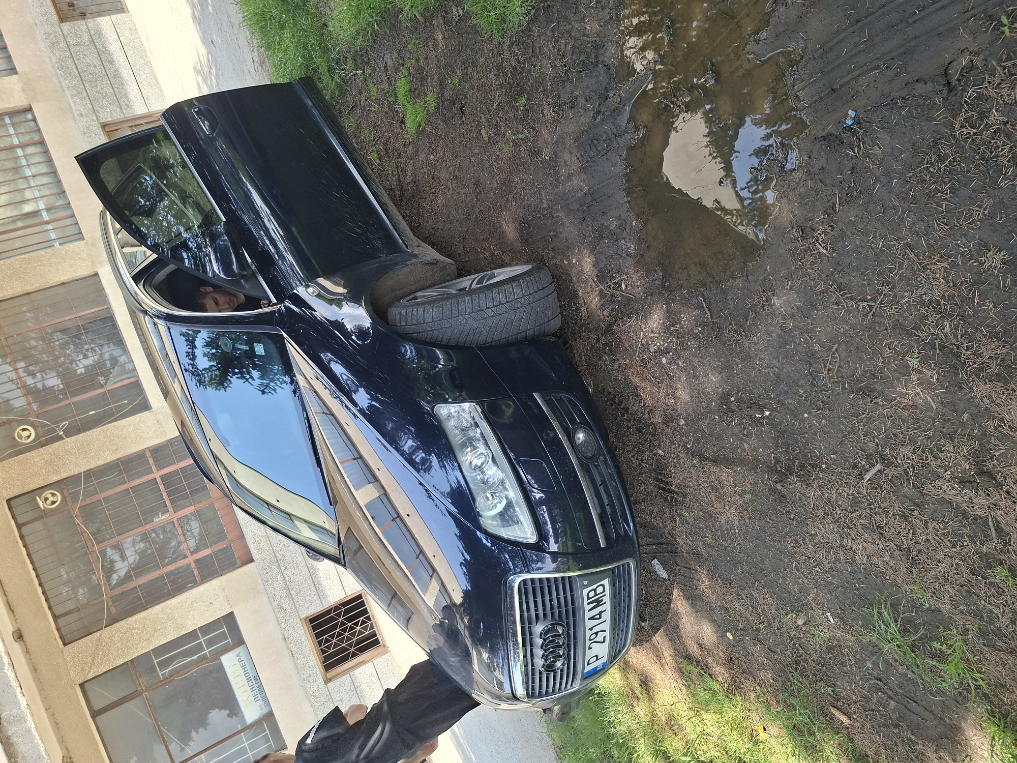 A dark-colored Audi A6 (C6 generation) station wagon, bearing Bulgarian license plate P 2914 MB (Ruse region), is depicted in Basarbovo, Bulgaria, parked partially off-road on a dirt and muddy surface next to a light-colored building. The car's front passenger wheel is situated in a distinct muddy area with a water puddle, showing tire tracks, while the rest of the vehicle is on firmer dirt mixed with grass and pine needles. The driver's side door is open.
Inside the vehicle, a male individual, appearing to be in his 20s or 30s with short dark hair and wearing a dark t-shirt, is seated in the driver's position. Another individual, also male and dressed in a dark t-shirt and dark pants, stands with his back mostly to the camera near the front driver's side of the car.
The background features a multi-story building with a light-colored facade. It contains several large, rectangular windows secured with metal grates. One prominent window displays a Cyrillic sign that translates to "Club for Pensioners." Smaller grated windows are also visible. Two round, fan-like fixtures are mounted high on the building's exterior. The scene is illuminated by bright daylight, casting visible shadows.