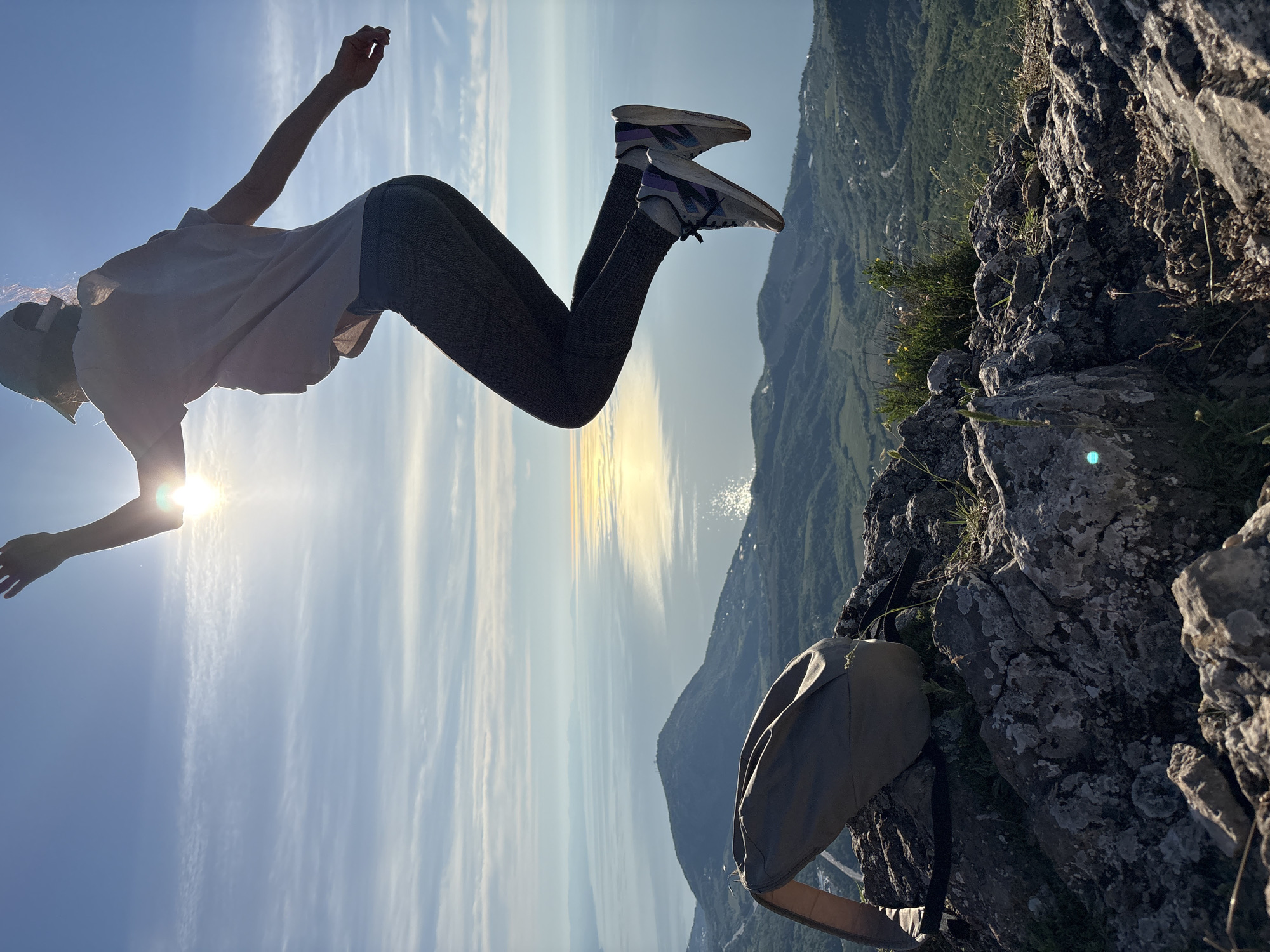 An outdoor, wide-angle shot captures a person mid-jump on a rocky mountain peak overlooking a vast body of water. The subject, silhouetted against the bright sun, is positioned in the upper center of the frame, facing slightly right and away from the camera. They wear a light-colored t-shirt, dark leggings, a baseball cap, and athletic shoes with purple and grey accents. Their arms are slightly raised, and legs bent, indicating upward motion.
In the immediate foreground, a rugged rock formation, partially illuminated by the sun, occupies the lower third of the image. A khaki or greyish-green backpack with an open top rests on these rocks to the left. Sparse green vegetation, including grass and small bushes, grows among the rocks.
Beyond the mountain peak, a sprawling landscape unfolds. Rolling green hills and mountains, dotted with small structures and roads, lead down to a large expanse of calm, blue water. The sun, located behind and to the upper right of the jumping person, creates a strong, shimmering reflection across the water's surface, extending towards the distant horizon. Wispy white clouds are visible in the bright blue sky above. The scene is illuminated by natural sunlight, casting the jumping figure in a partial silhouette with a prominent lens flare around the sun. The location is Unknown, Unknown.