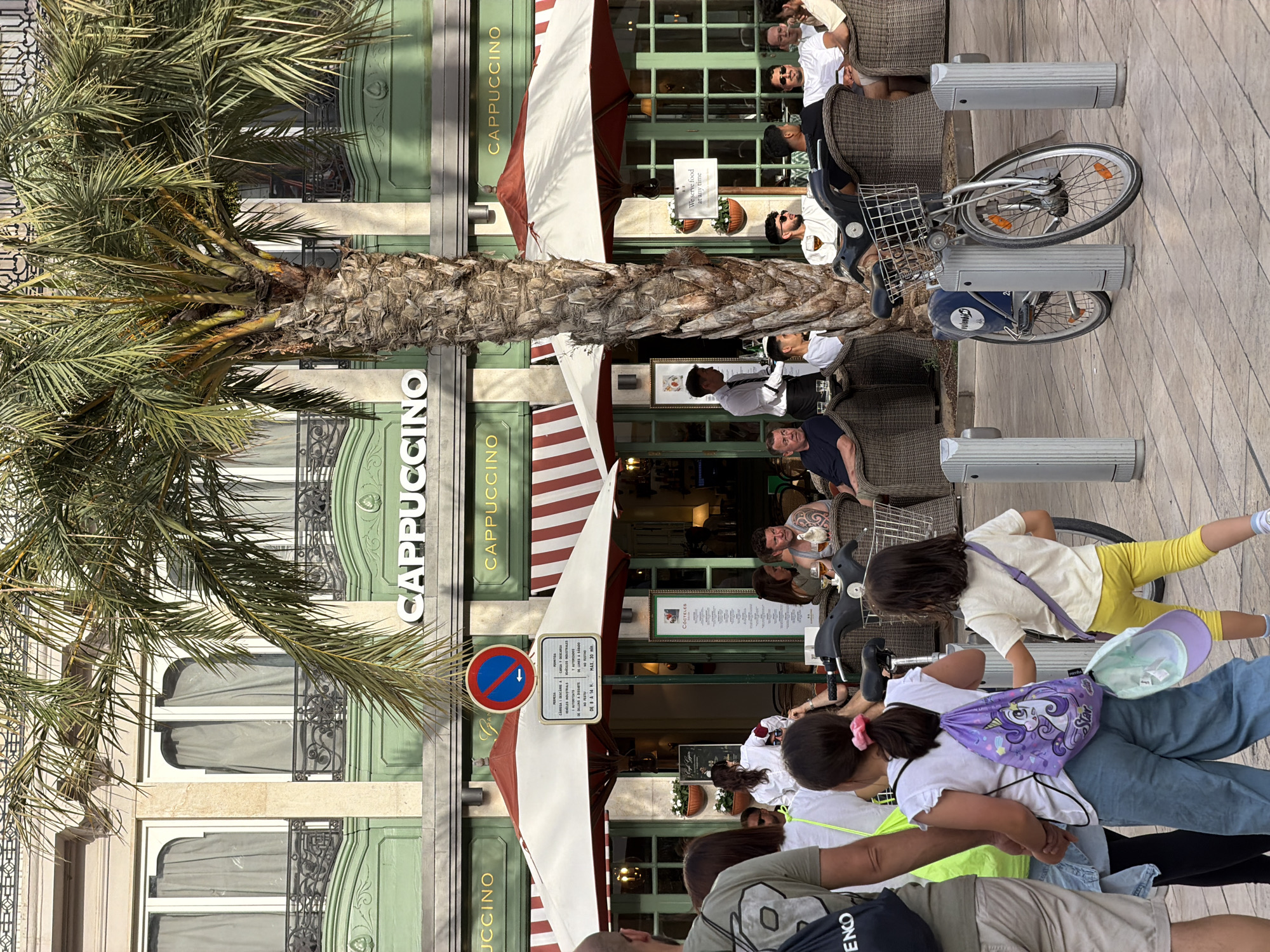 An outdoor seating area of a cafe called "Cappuccino" is shown on a sunny day, likely in a European city due to the architecture and the presence of a Vélo'o bike share station. In the foreground, a person wearing a black backpack and grey shorts walks from the left. Behind them, two young girls are walking away from the camera, one in blue jeans and a white shirt, carrying a purple unicorn backpack, and the other in yellow pants and a white t-shirt. A bicycle from the Vélo'o bike share is parked in front of the cafe's outdoor seating. In the middle ground, patrons are seated at outdoor tables under large umbrellas, some eating and drinking. A waiter in a white shirt and black tie is visible serving customers. In the background, the "Cappuccino" cafe has large green-trimmed windows and a sign with the cafe's name. A prominent palm tree stands in front of the building. Several other people are seated at tables, interacting with each other and enjoying their meals. A blue and red sign with an arrow and a crossed-out circle indicates a prohibition of some sort.