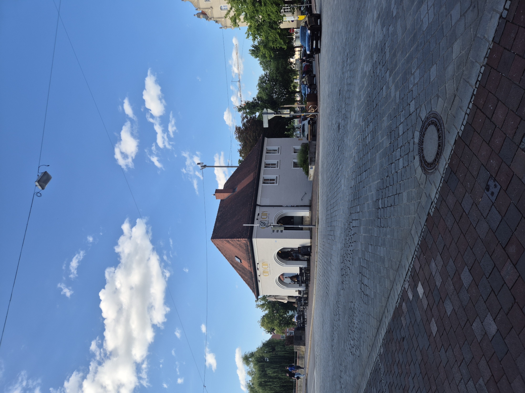 The image shows a street scene in Landshut, Germany. In the foreground, a cobblestone street is visible, with a manhole cover in the center. To the right, a black SUV is driving on the street. In the middle ground, a light-colored building with a red tile roof houses a cafe with outdoor seating. Several people are seated at tables outside the cafe, and others are walking in the background. A large tree with green foliage is on the left side of the building. The sky is blue with scattered white clouds. In the distance, there are more buildings and trees, and a construction crane is visible. The image appears to have been taken on a sunny day.