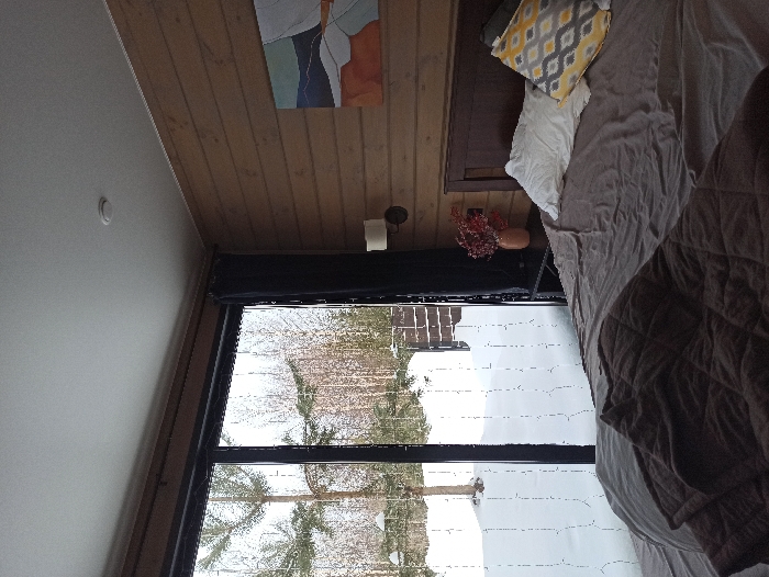 This image depicts the interior of a bedroom overlooking a snow-covered exterior in Nizhneye Zadolgoye, Russia. The room features horizontal light wood paneling on the walls and a white ceiling with a circular fixture. A large, dark-framed, floor-to-ceiling glass window or sliding door occupies the left side, providing a view of the outside. Dark curtains are partially drawn to the right of the window.
A large bed with a dark wooden headboard is central to the right portion of the image. The bed is covered with a grey duvet or sheet, and a dark brown quilted blanket is folded at the foot. Two pillows rest against the headboard: one white, and one with a yellow, grey, and white geometric pattern. To the right of the bed is a dark-colored nightstand. On the nightstand, a terracotta-colored vase holds a bouquet of reddish-orange artificial berries or foliage. Above the nightstand, a wall-mounted sconce with a cream-colored shade is affixed to the wooden wall. A rectangular abstract painting, featuring blues, grays, oranges, and whites, hangs on the wooden wall above the bed's headboard.
Through the window, the exterior ground is covered in a deep layer of snow. Bare deciduous trees and at least one coniferous tree are visible. A dark fence or partial structure is present in the background, further obscured by the snowy landscape. No individuals are depicted, and no specific actions or interactions are occurring.