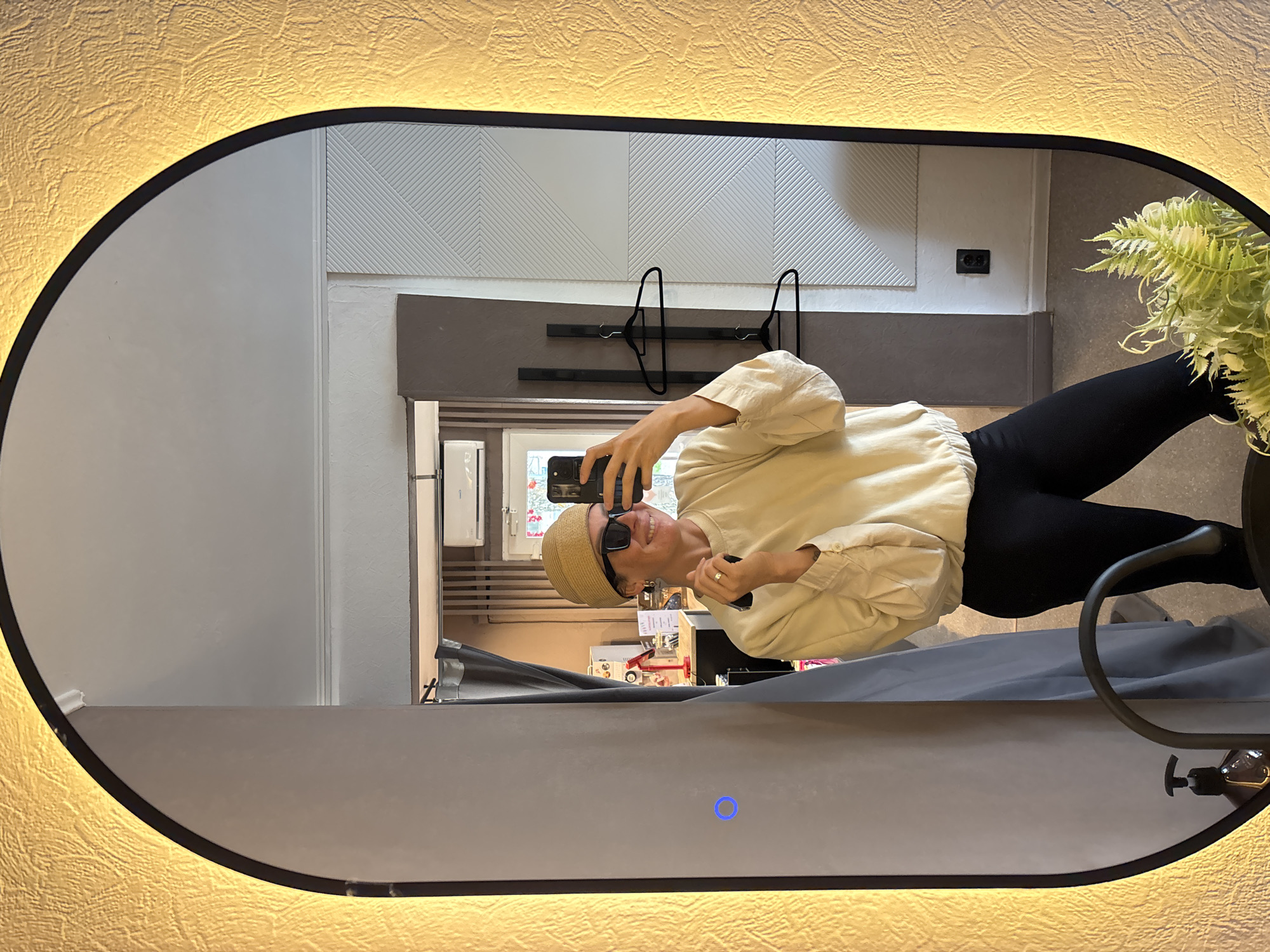 An augmented reality mirror, with a warm LED light and a textured yellow frame, reflects a woman in her late 20s or early 30s. She is wearing a tan beanie, black sunglasses, and a cream-colored sweatshirt with black leggings. She is holding a black smartphone in her right hand, taking a selfie, and a black compact object in her left hand. The mirror shows a room with a gray curtain, a white patterned wall, and a black electrical outlet. A plant with green leaves is visible in the foreground. The woman is smiling, and her left hand is resting on her stomach. This image was taken in Chaikovsky, Russia.