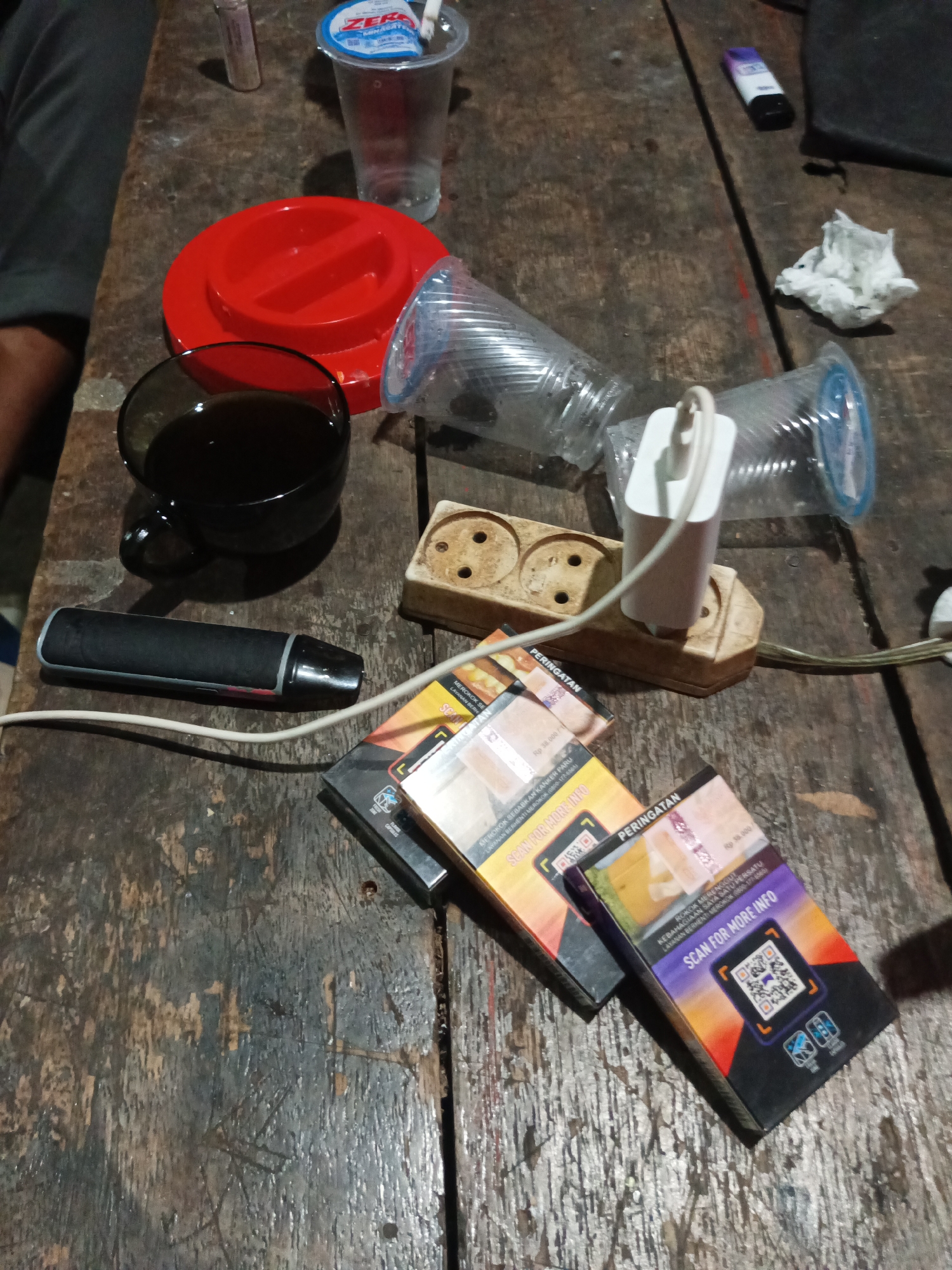 The image shows a close-up, top-down view of a rustic wooden table with various objects scattered on it. In the foreground, two packs of cigarettes are prominently displayed, featuring prominent warning labels and QR codes. To the left of the cigarette packs, a black electronic cigarette device rests on the table. Behind the cigarettes, a white power strip with two electrical outlets is visible, with a white phone charger plugged into one of the outlets, its cord extending towards the bottom right. A black coffee cup filled with a dark liquid sits to the left of the power strip. A large red lid, possibly for a container, is positioned behind the coffee cup. To the right of the red lid, two clear plastic cups are present, one lying on its side and the other upright. Further back, a crumpled white tissue is visible. The background is out of focus but suggests an indoor setting with dimly lit ambiance. The overall scene depicts a casual arrangement of personal items and consumables on a worn wooden surface. There is no visible indication of the city of Tumampua, Indonesia.