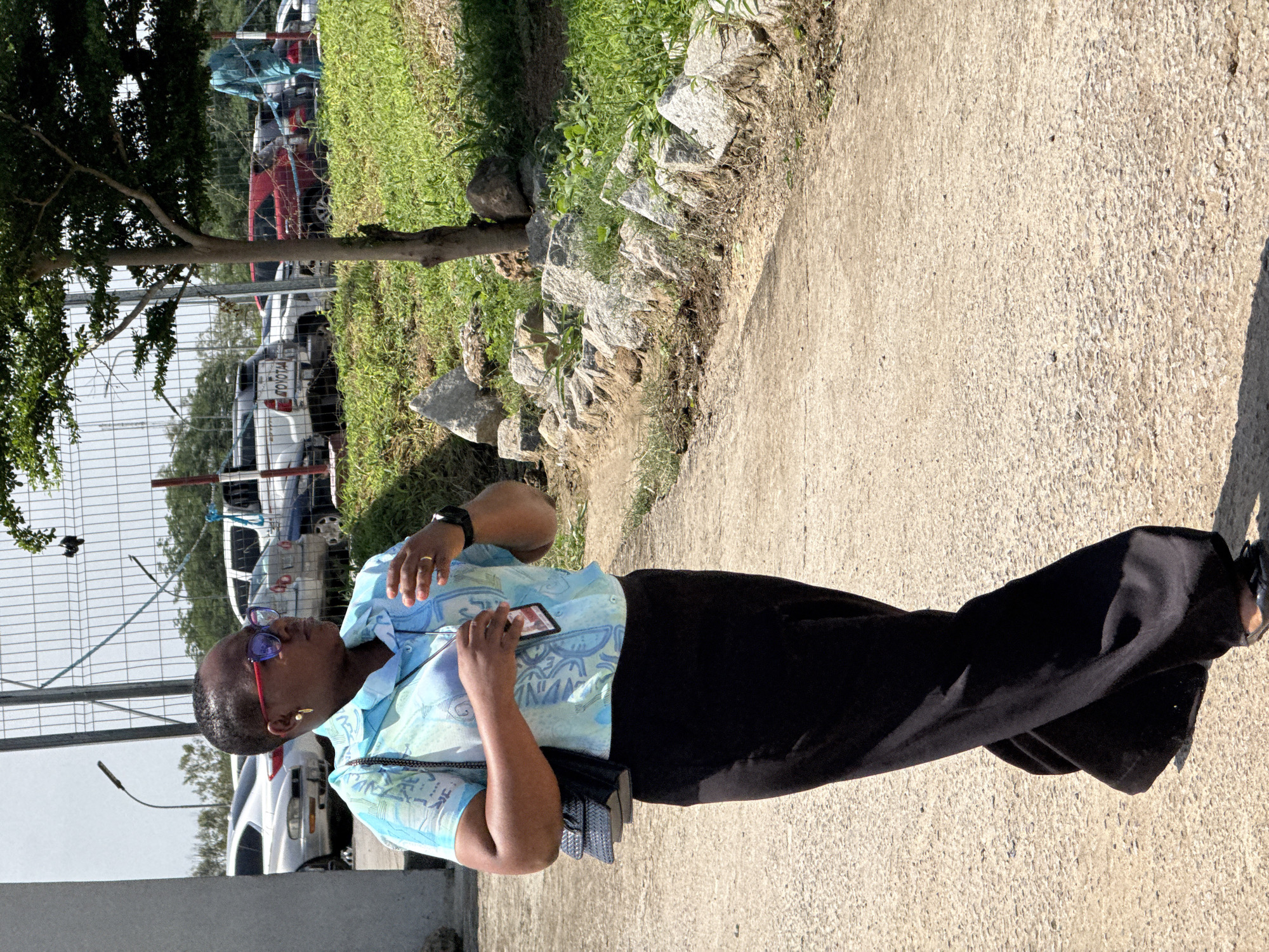 In Gwaska, Nigeria, on a bright day, a Black woman wearing red-rimmed glasses, a light blue and green patterned short-sleeved shirt, and a long black skirt walks on a paved path. She carries a black quilted purse on her left shoulder. Her right hand is raised, with her fingers extended as if gesturing or adjusting something near her chest. A lanyard with an ID card is visible around her neck. The path is lined with rocks and green grass on the right, and a concrete wall and a fence with vehicles in the background on the left. A small tree with dark green leaves stands between the path and the fence. The scene appears to be an outdoor area, possibly a parking lot or a facility.
