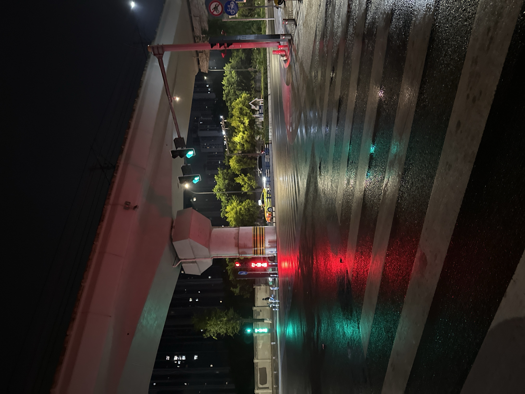 This image depicts a nighttime urban street scene in the Chongqing High-tech Zone, China, following a recent rainfall, evidenced by the wet, reflective road surface. An elevated concrete overpass structure, supported by a large, multi-faceted pillar with yellow and black hazard stripes, dominates the upper portion of the frame. Below the overpass, a busy intersection is illuminated by various light sources.
On the left, a set of two vertical green pedestrian traffic lights stands on a pole, and further right, suspended beneath the overpass, are two green vehicle traffic lights. A third traffic light pole on the right side of the image displays red pedestrian lights. The wet asphalt road exhibits strong, vivid reflections of these lights, with a prominent green hue reflecting from the left side onto the crosswalk, and an equally strong red reflection dominating the right side of the crosswalk and road.
A multi-lane zebra crossing is visible in the foreground, its white stripes reflecting ambient light while the dark asphalt reflects the colored traffic signals. A person on a white scooter or small motorbike is stopped near the center-left of the intersection, positioned behind the crosswalk. In the mid-ground, a yellow taxi is visible on the far right side of the street, and a dark sedan is further back to its left.
The background features numerous tall, multi-story buildings with illuminated windows, indicating a densely populated area. Lush green trees and shrubs line the streets and a central median strip, some appearing yellowish from artificial illumination. Various street poles, barriers, and traffic signs, including a "no motor vehicles" and "no bicycles" sign, are present on the right side. The sky is dark, showing faint ambient light sources in the distance.