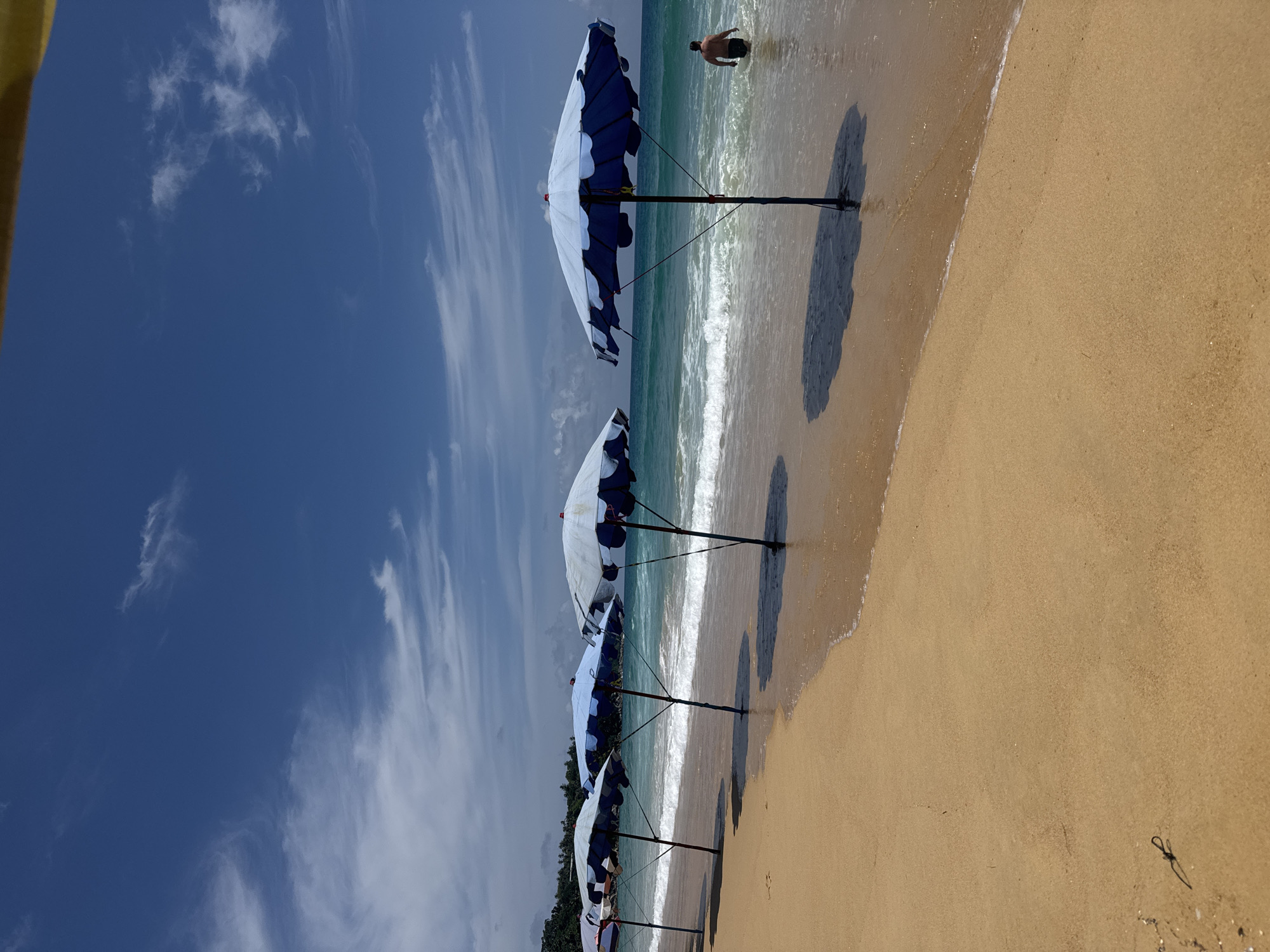 A row of blue and white beach umbrellas are lined up on a sandy beach in Karon, Thailand. The umbrellas are set up on the sand, with some casting shadows on the wet sand where the waves are gently lapping. The ocean is a vibrant turquoise color, and the sky above is a clear blue with scattered white clouds. In the background, a lone man is standing in the shallow water, facing away from the camera. The beach appears to be quiet, with no other people visible in the immediate vicinity. The overall scene depicts a serene beach environment, likely during the daytime given the bright sunlight.