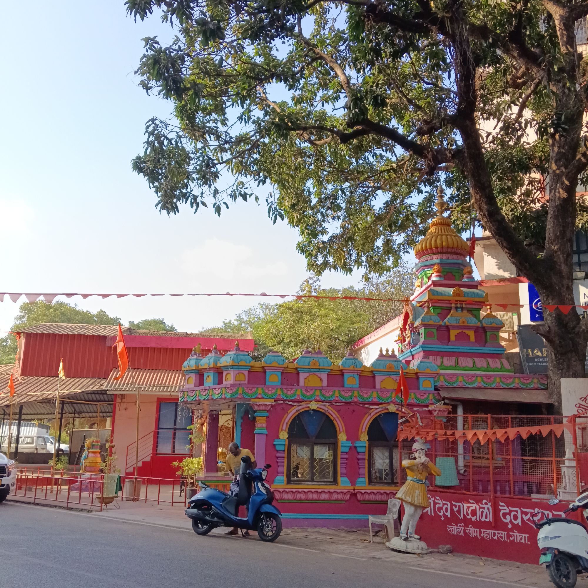 The image shows a colorful Hindu temple on a street in Mapusa, Goa, India. The temple is painted in bright pink, blue, green, and yellow hues, with intricate designs and statues. A person is standing next to a blue scooter parked in front of the temple. A white statue of a man stands near the temple wall, which is painted red and features the temple's name in Devanagari script: "श्रीदेव राष्ट्रोळी देवस्थान, खोर्ली सीम, म्हापसा, गोवा." A row of pennants hangs above the temple and adjacent buildings. A large tree partially obscures the view of the temple's upper section. A road runs in front of the temple. The image has been taken during daytime under the blue sky.