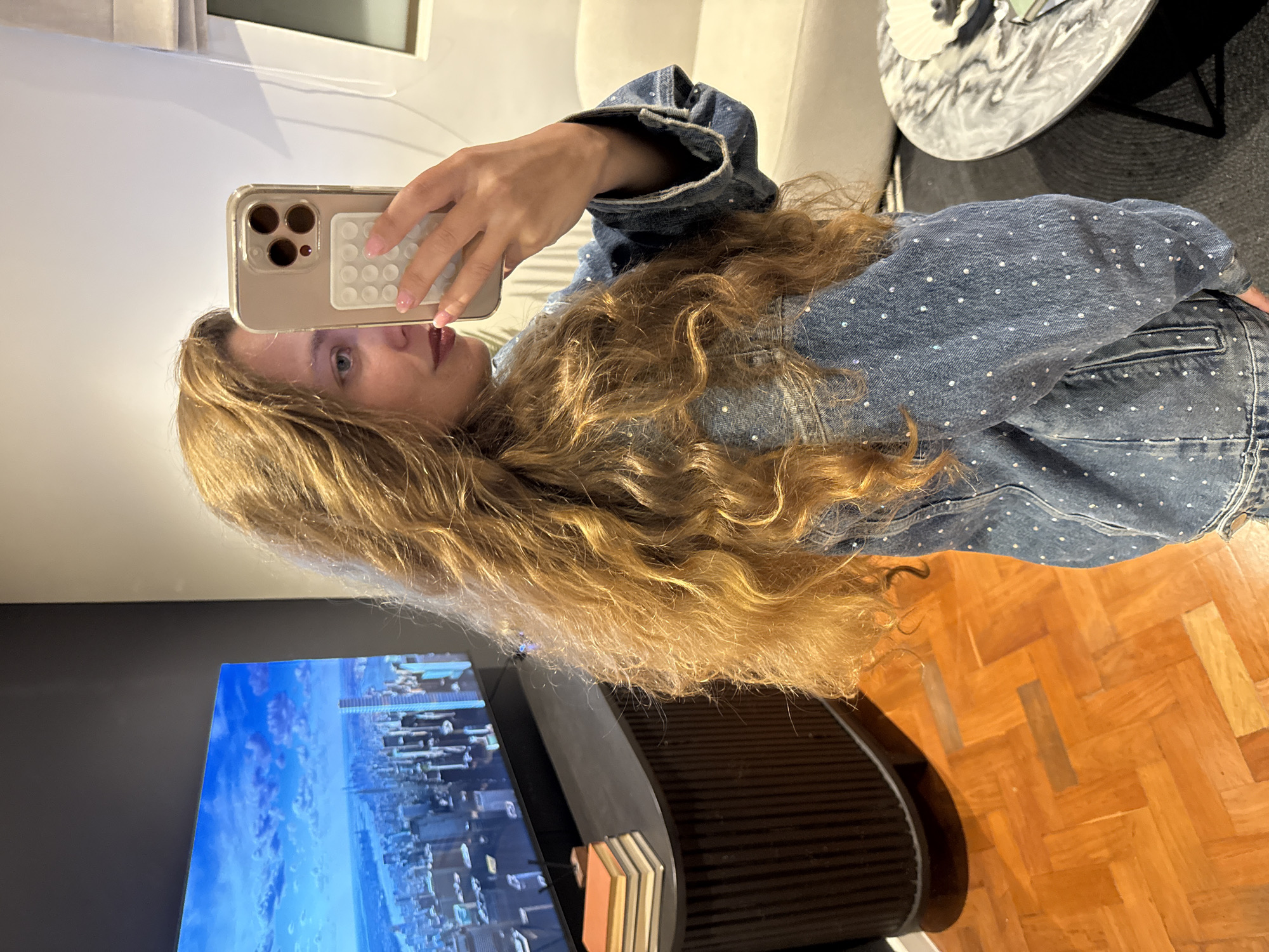 A young woman with long, wavy blonde hair is taking a selfie with a gold iPhone. She is wearing a denim jacket and jeans studded with small, shiny embellishments. In the background is a modern living room with hardwood floors. A television displays a cityscape that resembles the city of Cape Town, South Africa, including a view of the coastline and skyscrapers. A black TV stand sits under the television, with a stack of books on one side. In the upper background, there is a white sofa and a marble side table.