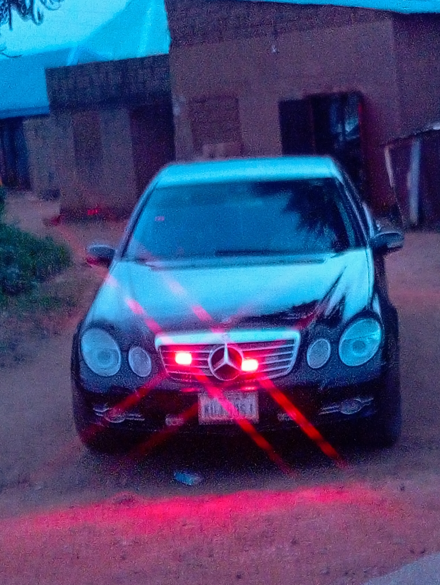 The image shows the front of a black Mercedes-Benz sedan parked on a dirt road. The car has its headlights and front grill illuminated with a reddish glow, suggesting the lights are on or reflecting from another source. The car's license plate, which appears to be "KDA 951", is visible on the front bumper. In the background, there is a building with a dark-colored facade, possibly a house or a commercial structure. The ground around the car is covered with loose dirt and gravel. The overall lighting suggests it might be dusk or nighttime. The scene is located in the city of Du, Nigeria, based on the provided context, though no specific landmarks within Du are identifiable from the image itself.