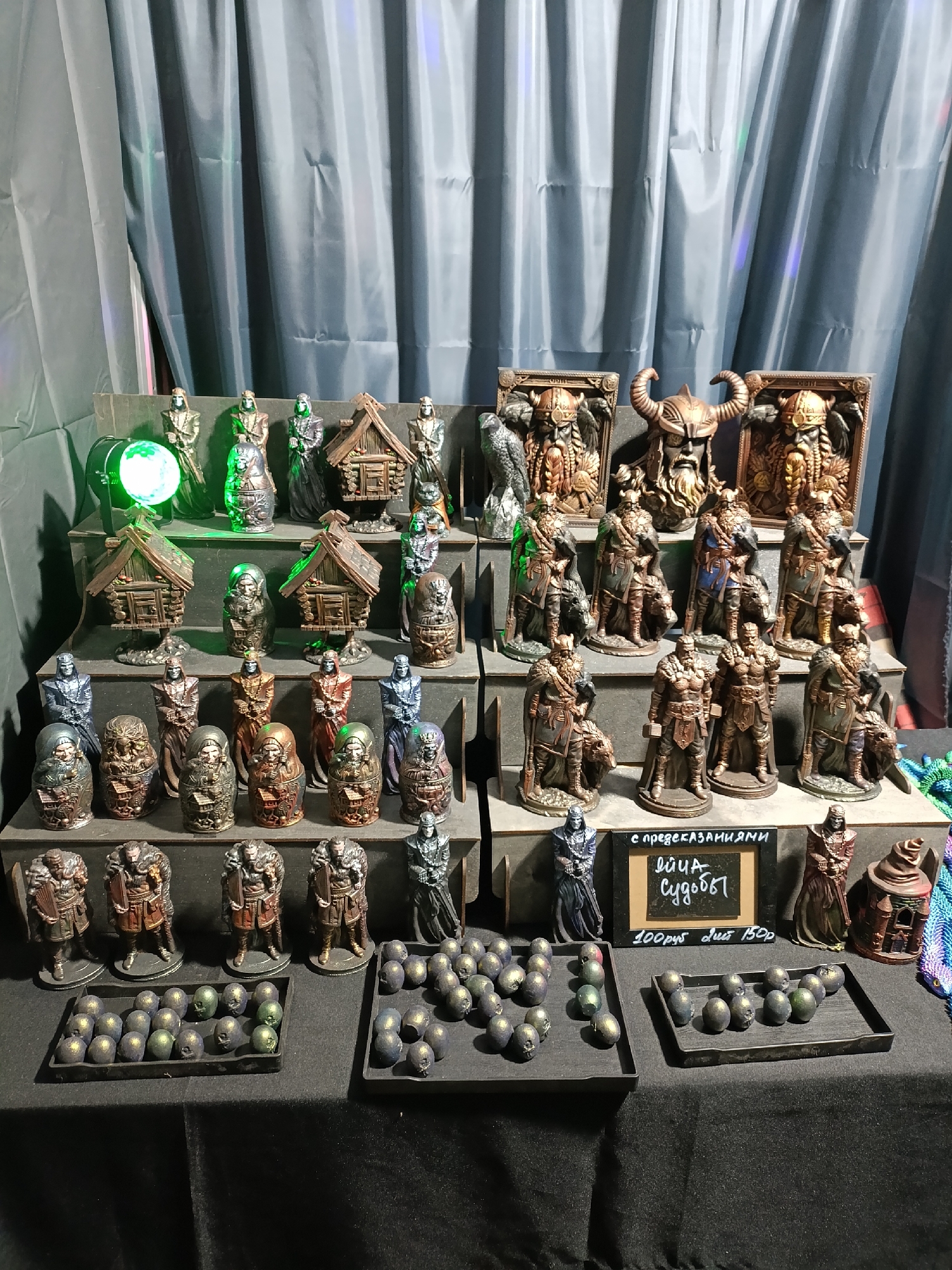 This image depicts a display of hand-painted miniature figurines and decorative items. The items are arranged on tiered shelves and on a table covered with a black cloth. The figurines appear to be of a fantasy or historical theme, featuring characters in armor, warrior-like figures with helmets and beards, and small, rustic structures resembling houses. Some figures are set against elaborate backgrounds depicting faces with helmets and horns. Scattered among the figurines are small, round objects, some appearing metallic and others with a greenish sheen. A small chalkboard sign with handwritten text in Russian is also visible, accompanied by a price. The background consists of a dark, pleated curtain, and to the left, a green light emanates from a spherical object. The overall setting suggests a stall or display at a market or event. This scene is presented as if it were located in the city of Lagunovo, Russia.