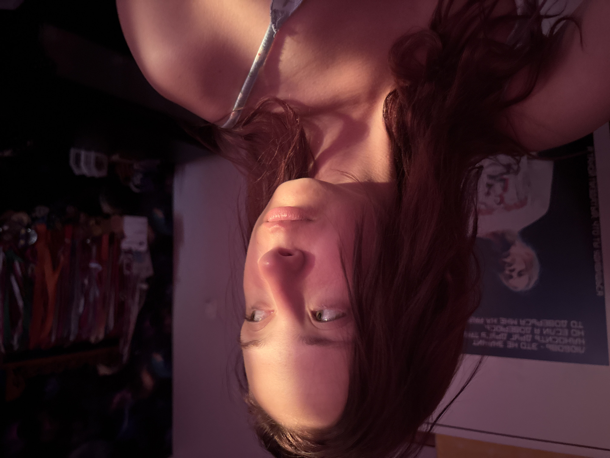 The image is an upside-down close-up of a woman's face. Her hair is reddish-brown. She has light skin and eyes. Her lips are closed. The background is blurred, but there appears to be a clothing rack on the left side and a poster with text on the right. The text on the poster is in Russian and partially visible. The location is in the city of Прикамский, Russia.