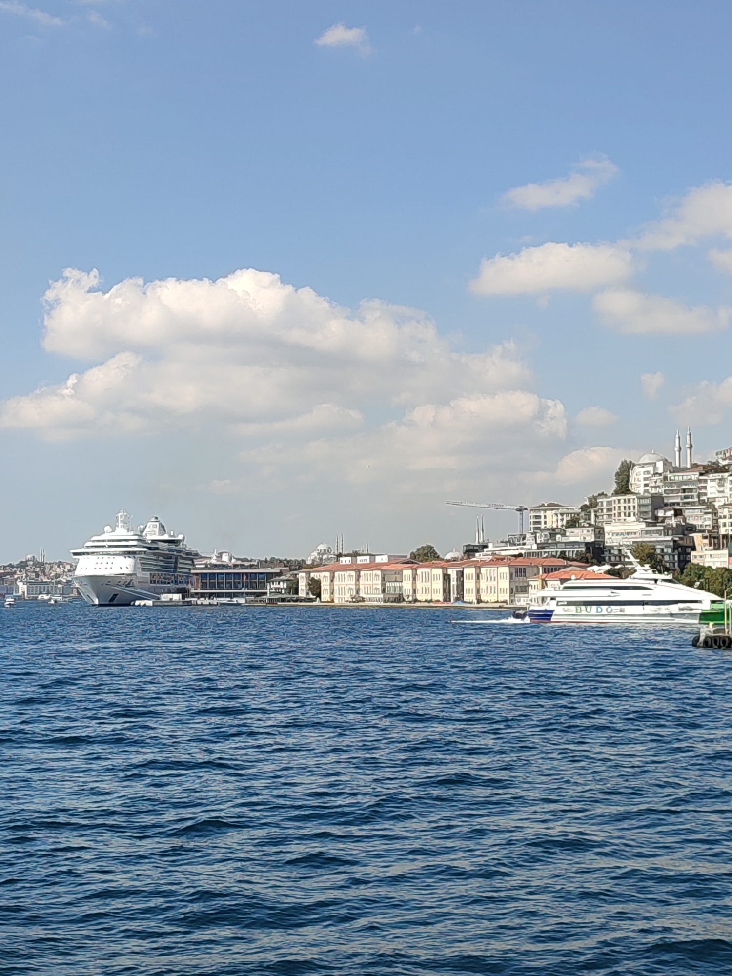 The image depicts a daytime view of the Bosphorus strait in Istanbul, Turkey, specifically showing a waterfront area likely in or near the Beyoğlu district. In the foreground, the deep blue water of the strait is textured with small waves. Moored along the waterfront is a large, white cruise ship, appearing to be docked at a terminal with a long, modern building behind it. To the right of the cruise ship, a sleek, white passenger ferry with the text "BUDO" visible on its side is in motion, leaving a white wake in the water. Further down the coastline, a dense collection of buildings, many with white facades and red-tiled roofs, ascend a hillside. Two minarets rise above the buildings, characteristic of mosques. In the distance, the skyline is punctuated by the silhouettes of other structures and cranes, suggesting ongoing construction or development. The sky above is a clear blue, with scattered white cumulus clouds. The overall scene conveys a sense of maritime activity and urban coastal life in Istanbul.