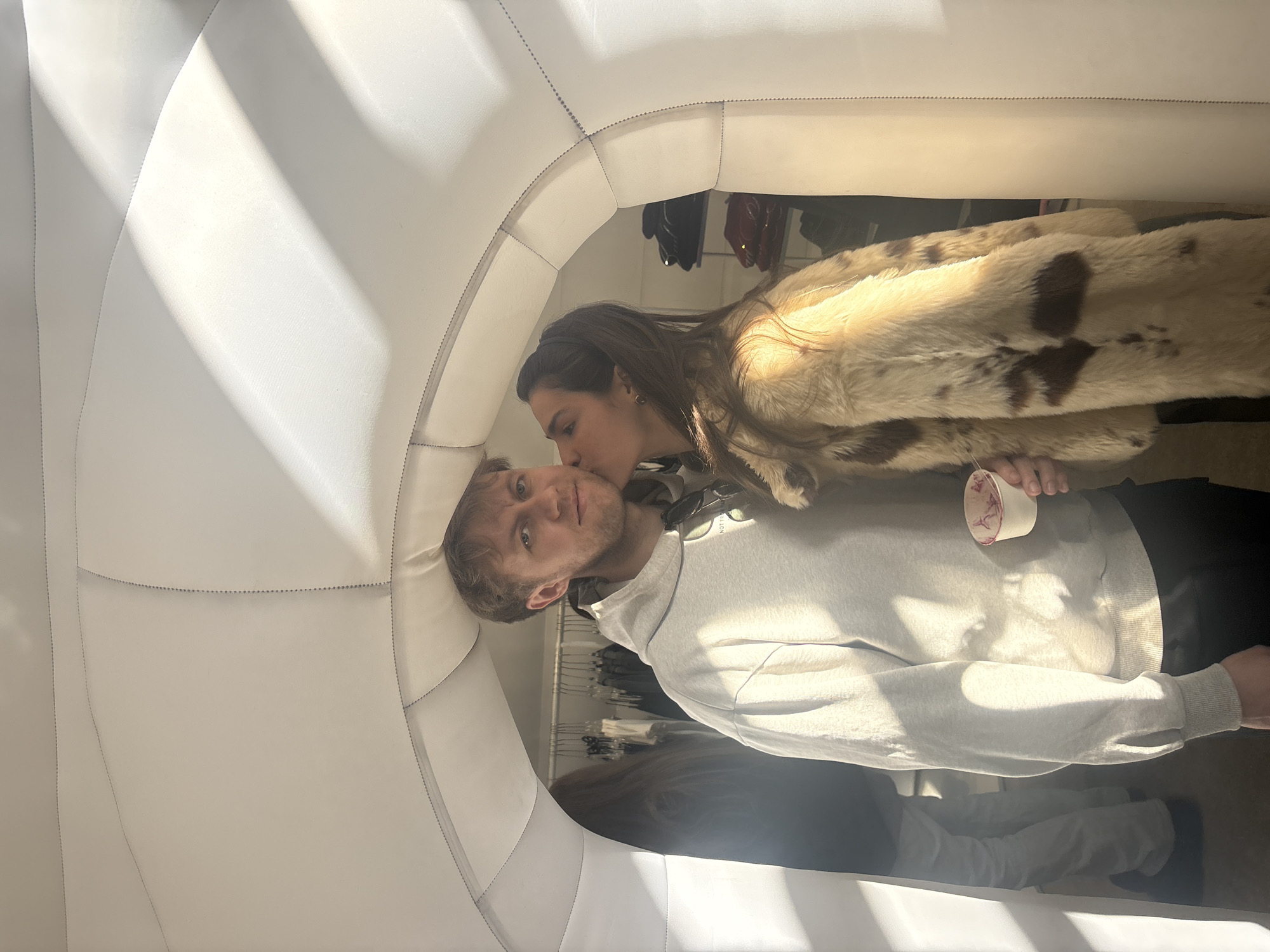 A woman kisses a man on the cheek inside a white, padded booth. The woman wears a fur coat and has long brown hair. The man wears a gray hoodie and holds a cup. Clothing racks are visible in the background. The location is possibly a store, but it is not in Moscow, Russia.