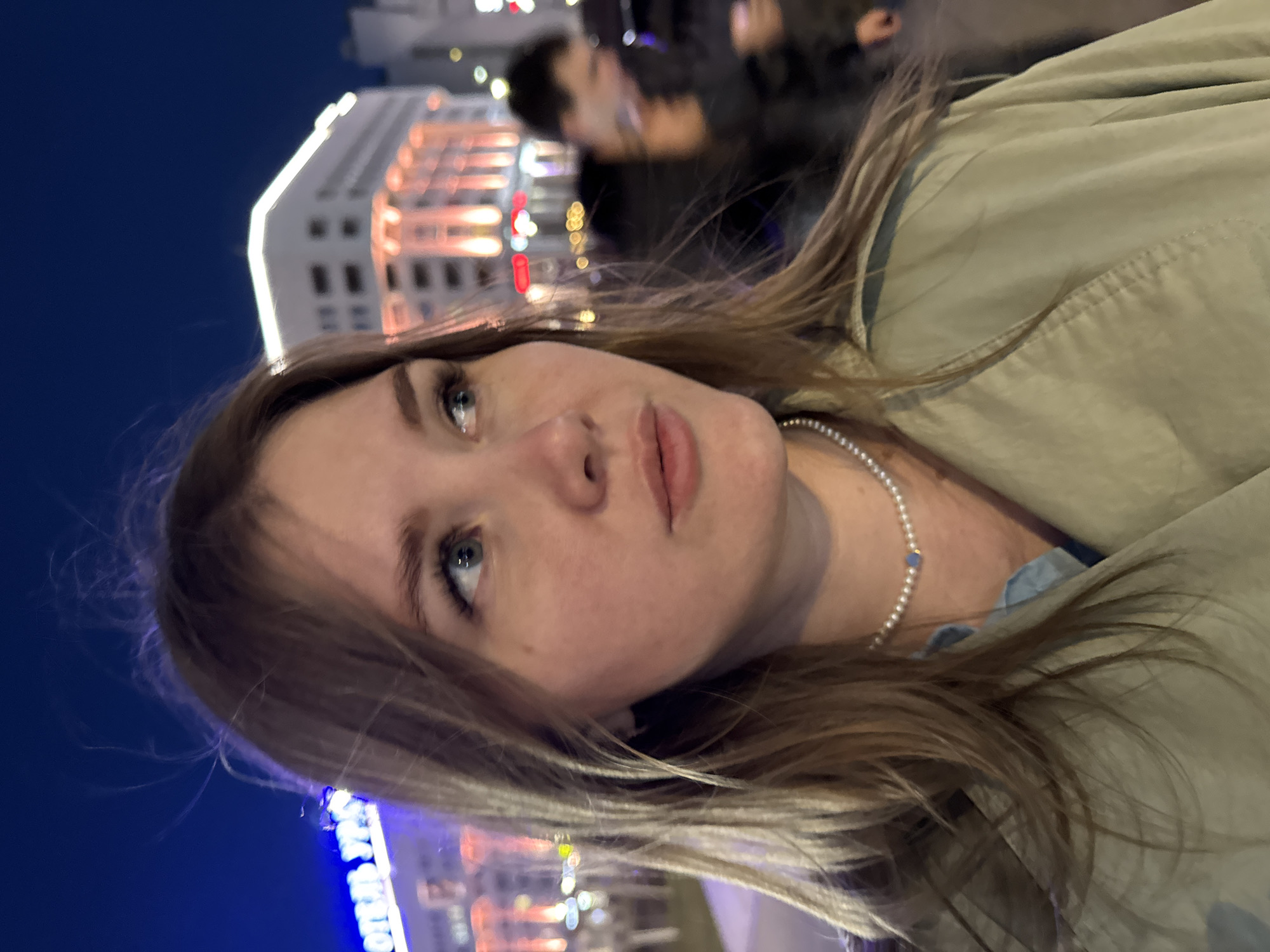 The photograph is an off-kilter, eye-level view of a woman with shoulder-length light brown hair and fair skin. She is wearing a light green jacket and a pearl necklace with a blue accent bead. The woman is looking upwards and to the left. Her expression is neutral.
In the blurred background, a building with illuminated windows is visible. Above the woman's head and behind her are other people in the background who are out of focus.
Based on the blurred sign at the bottom of the image, the location is in the city of Perm, Russia.