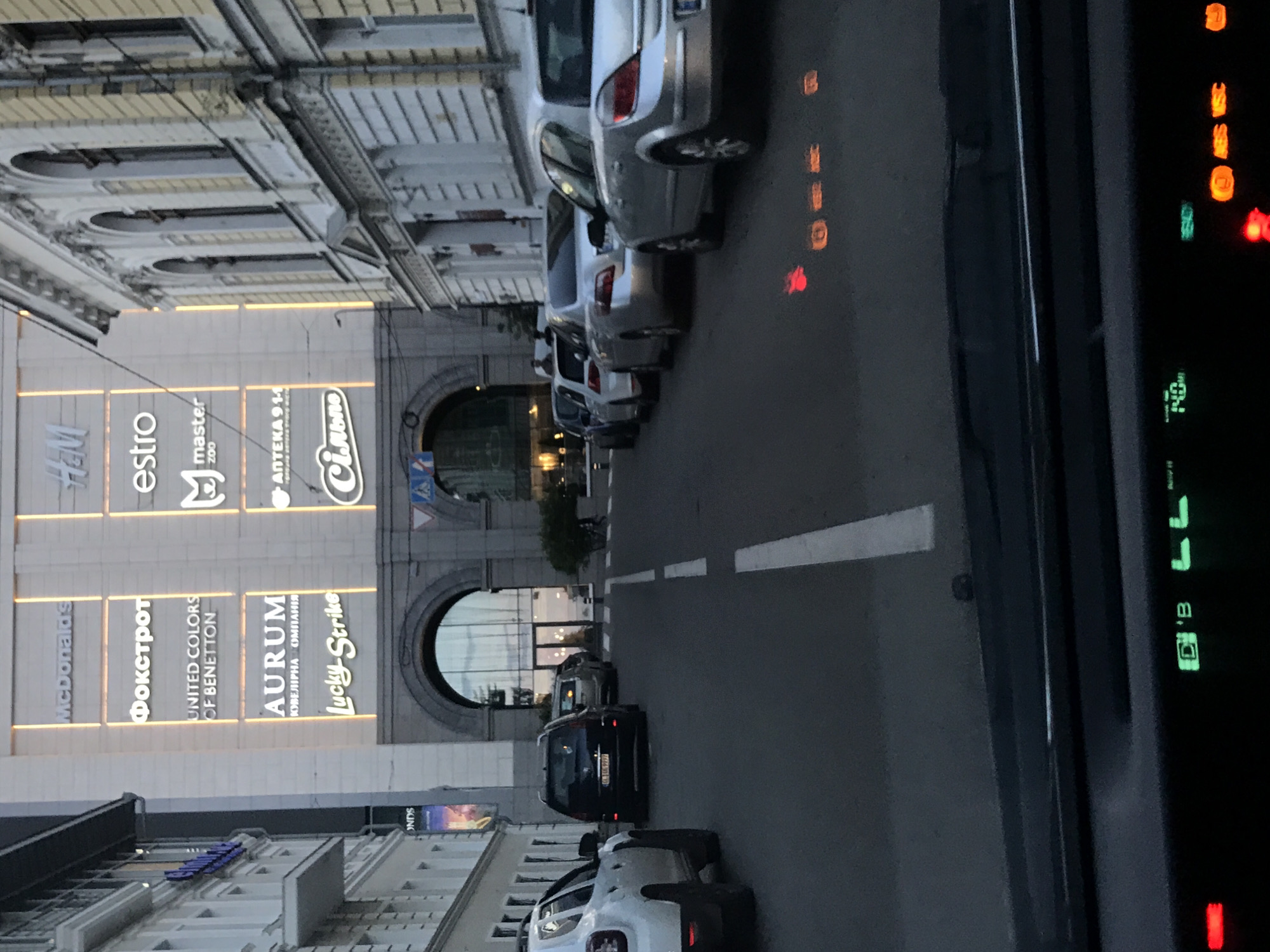 The image depicts a street scene in the city of Unknown, Unknown. On the left side of the street, there is a large building adorned with illuminated signs for various businesses, including McDonald's, Фокстрот, United Colors of Benetton, AURUM, Lucky Strike, Сільпо, H&M, estro, master zoo, and АПТЕКА 9-1-1. Arched entrances are visible within the building's facade. Along the roadside, several cars are parked in a row, facing the same direction. The road is marked with a white dividing line. The photograph appears to have been taken from inside a vehicle, as indicated by the dashboard and various indicator lights visible in the foreground on the right side of the image.