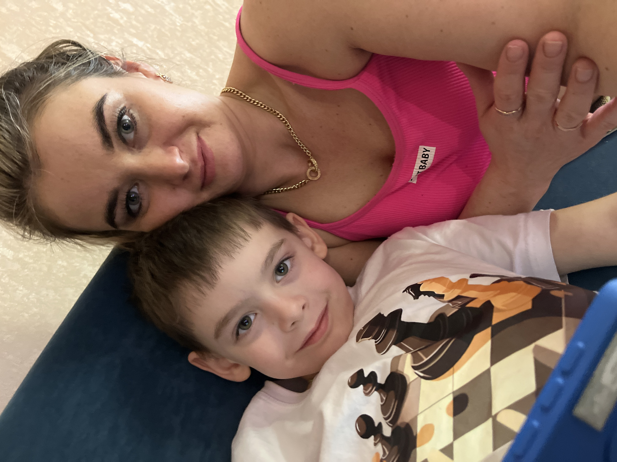 This image captures an indoor scene in Chaikovsky, Russia, depicting an adult female and a young boy in a close-up, chest-up view. The female, positioned on the right, has light hair, light eyes, and is wearing a ribbed bright pink tank top with a "BABY" label on the chest. She wears a gold-colored chain necklace with a circular accent, a small gold earring in her right ear, and two rings on the ring and middle fingers of her visible left hand. Her left arm is partially visible, appearing to be around or beside the boy. She is looking directly at the camera with a slight smile. The boy, positioned on the left, has short light-brown hair, light-colored eyes, and is wearing a white t-shirt featuring a graphic of dark chess pieces on a chessboard pattern. He is also looking directly at the camera with a slight smile. A dark blue upholstered surface, consistent with a sofa or large pillow, is visible behind the boy. A portion of a blue-cased electronic device is visible in the bottom-left corner, near the boy. The background consists of a light-colored, textured wall.