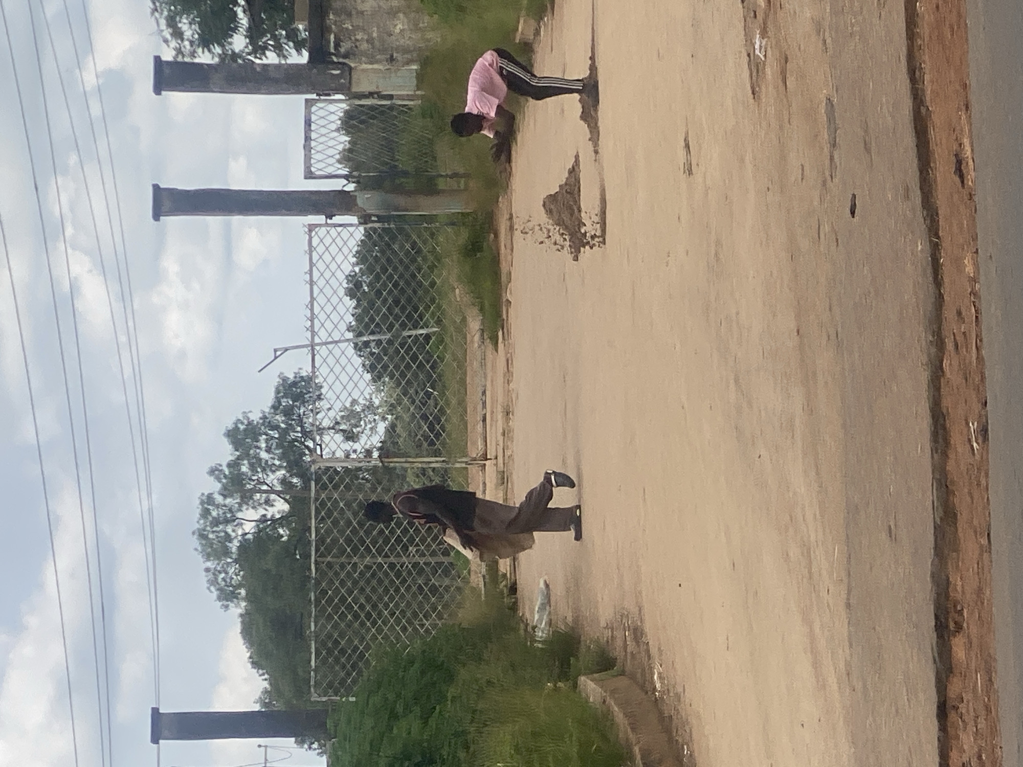 Two individuals are in a dirt road in Rafin Par, Nigeria. On the left, a person wearing a dark sleeveless shirt and grey pants walks away from the camera, carrying a large, light-colored sack. On the right, a person in a pink t-shirt and black pants with white stripes is bent over, scattering a pile of dirt into the air. In the background, there is a chain-link fence, large pillar-like structures, and trees. The sky is overcast.