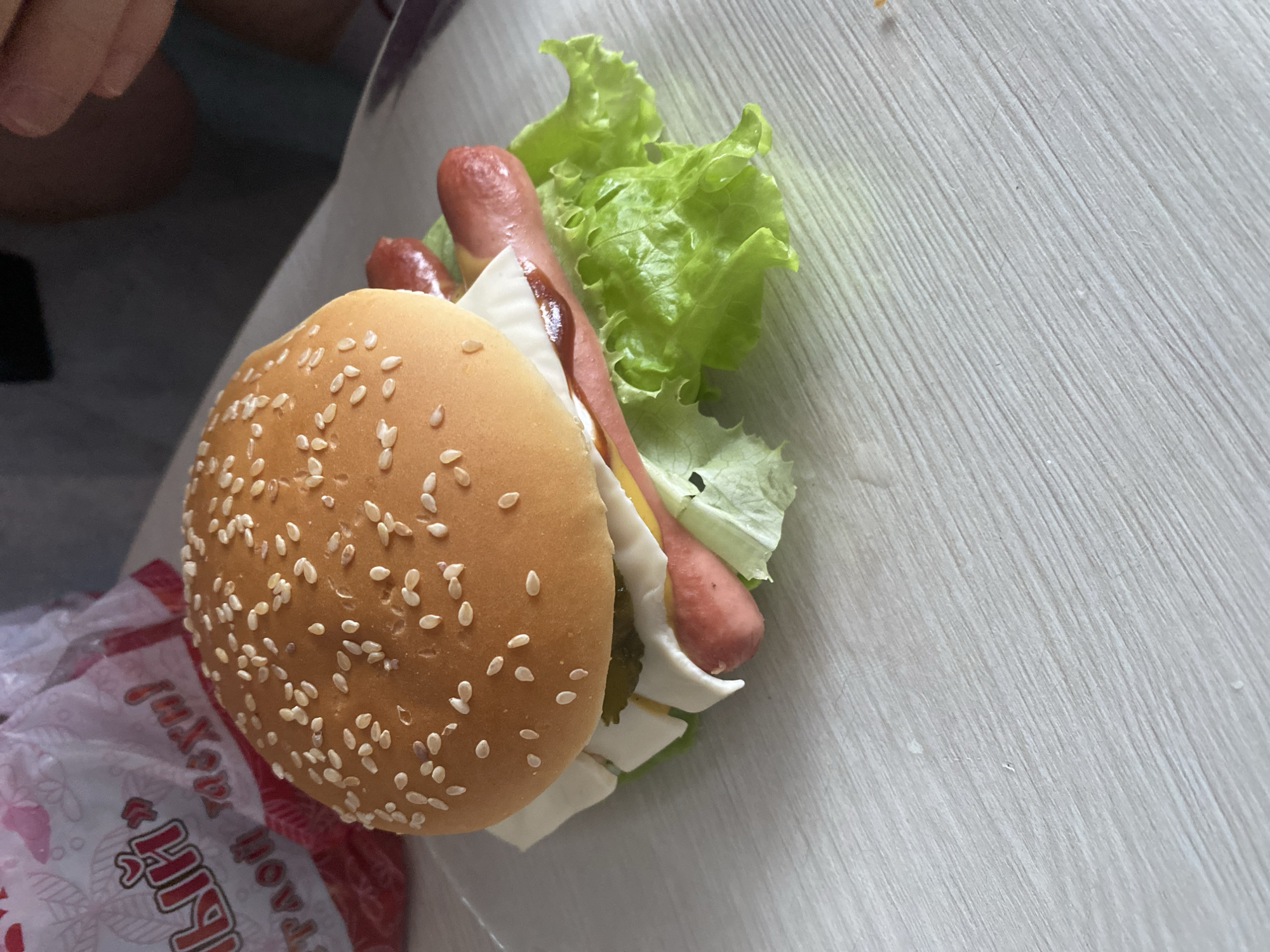 A close-up, overhead shot shows a partially assembled hot dog in a sesame seed bun. The hot dog appears to contain lettuce, cheese, pickles, mustard, and a dark sauce, possibly ketchup or barbecue sauce. The hot dog sits on a white surface, possibly a table. A partially visible red and white wrapper is near the bun. The location of the image is unspecified. The city of Izhevsk, Russia, is also unspecified.