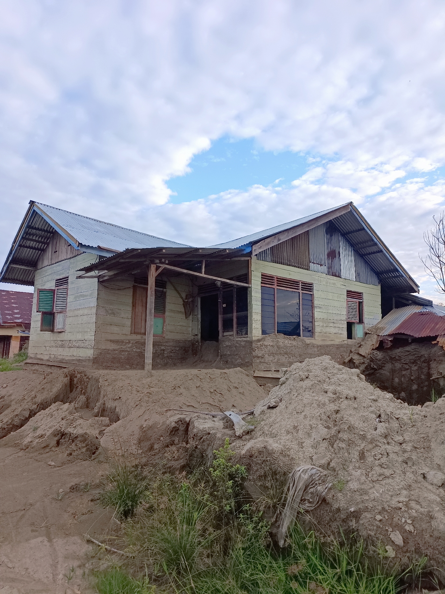 The image shows a light-colored wooden house in Meunasah Raya, Indonesia. The house has a corrugated metal roof and several windows. The ground around the house is disturbed, with piles of dirt and trenches, suggesting some sort of excavation or construction work is in progress. The sky is partly cloudy. There are no people visible in the image.