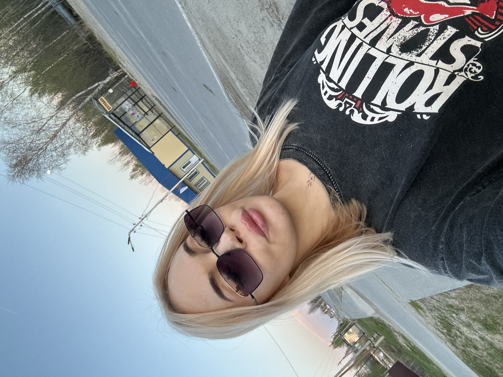 A woman with blonde hair and sunglasses is posing in an outdoor setting. She's wearing a dark sweatshirt with a "Rolling Stones" logo. The background includes a blue and beige bus stop structure with a sign in Russian that indicates "остановка" in Высокий, Russia, as well as a grey road, green vegetation, trees, power lines, and a sky transitioning from blue to a light pink-orange at the horizon.