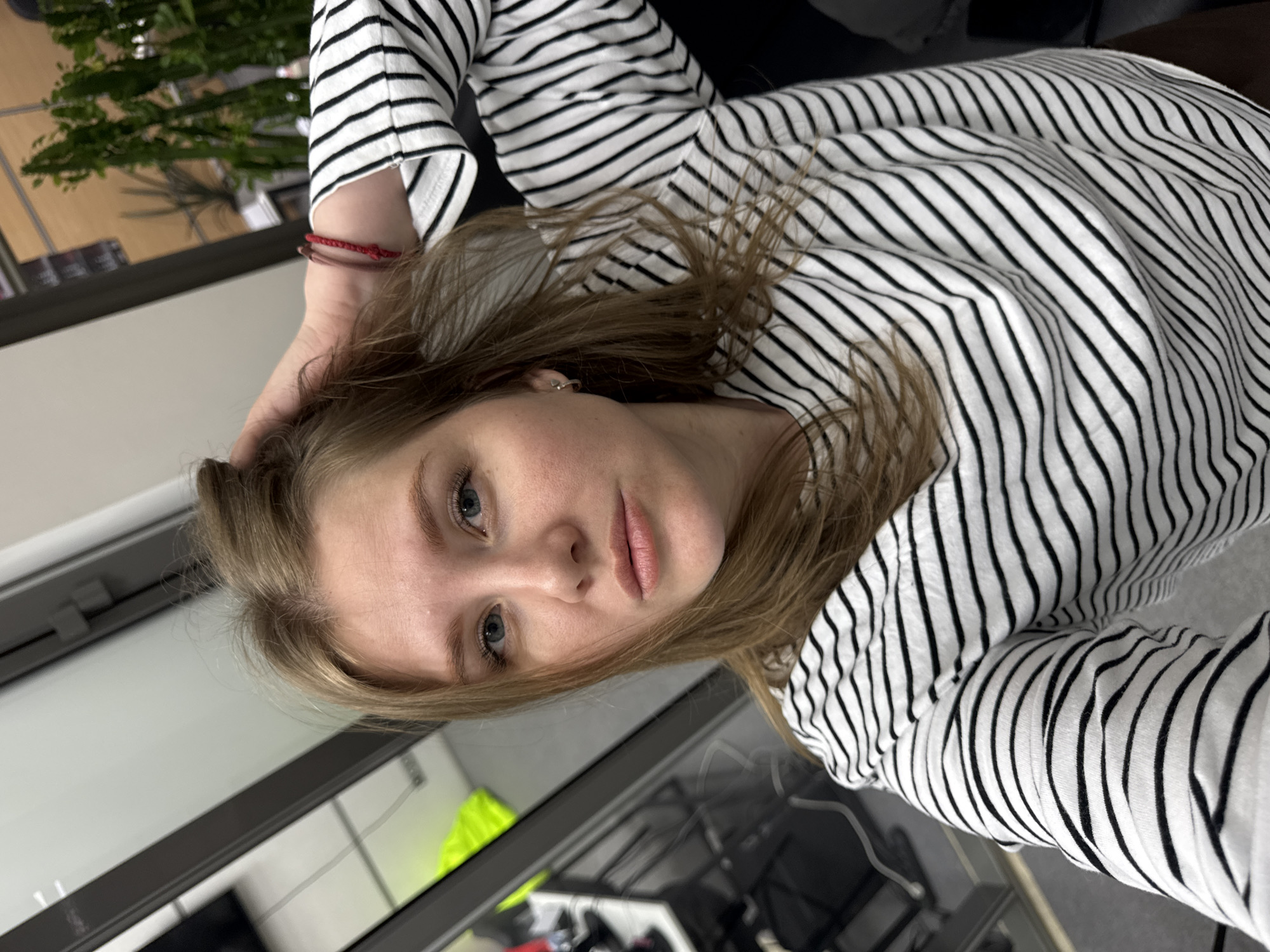 A young woman with fair skin and light brown hair, wearing a black and white striped long-sleeved shirt, is seen in a medium shot. Her head is tilted, and she is looking directly at the camera. Her left hand is raised to her hair, and two red bracelets are visible on her left wrist. The background includes a window with a grey frame, a green plant, and items on a desk below. The location cannot be determined as being in Perm, Russia.