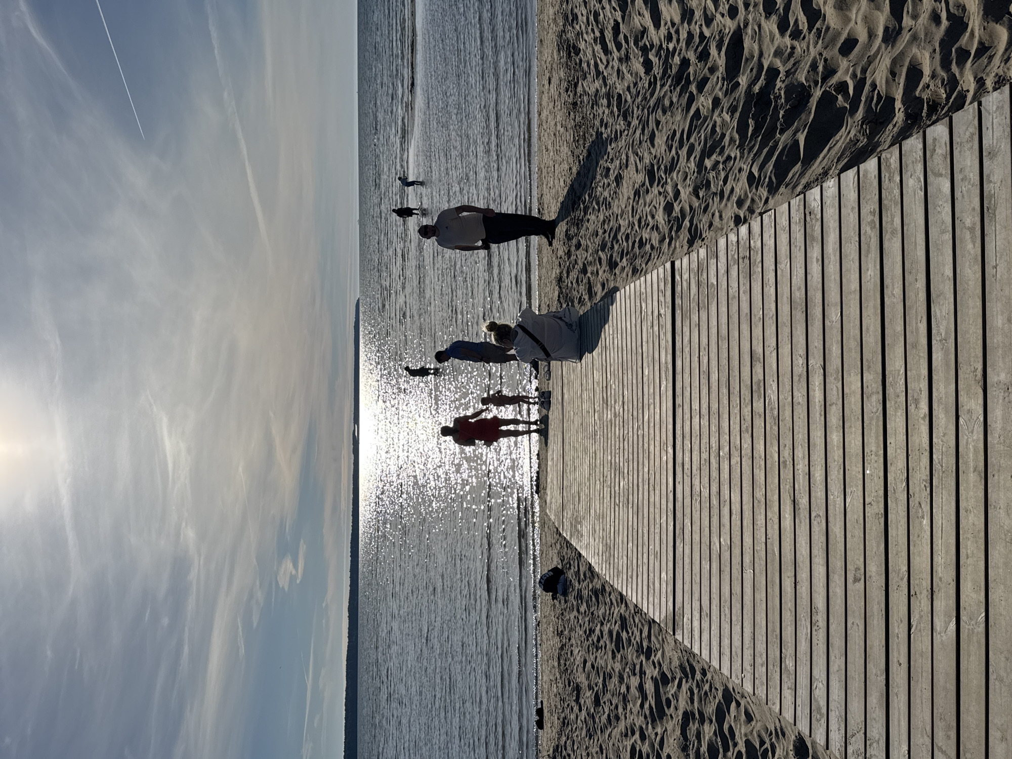 The image depicts a sun-drenched beach scene with a wooden boardwalk leading towards the water. Silhouetted figures of people are scattered across the beach and wading in the shimmering water, with a prominent group near the boardwalk. The sun is high in the sky, casting a bright glare on the water and creating distinct shadows on the sand. The background shows a distant treeline and a hazy horizon under a sky with wispy clouds and a contrail from an airplane. This scene is located at Pirita Beach in Tallinn, Estonia.