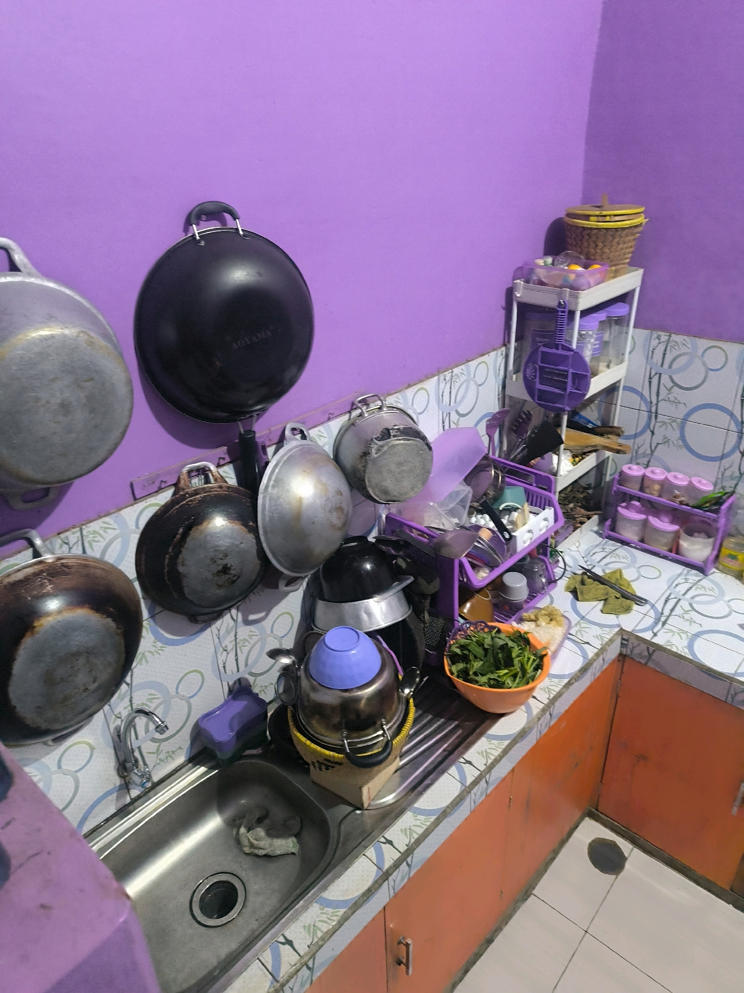 The image shows a cluttered kitchen counter and wall. On the purple wall, several pots and pans are hanging. The counter is tiled with a blue and white pattern. To the left, there is a stainless steel sink with a faucet. To the right of the sink, there are several kitchen items, including a purple rack with utensils, a basket of green leafy vegetables, and a tea kettle with a blue lid. Further to the right, there is a white shelf unit holding various containers and a woven basket with a yellow lid. The cabinets below the counter are orange. The overall impression is of a functional but messy kitchen environment. There are no people visible in the image. This kitchen is located in Cilengkrang, Indonesia.