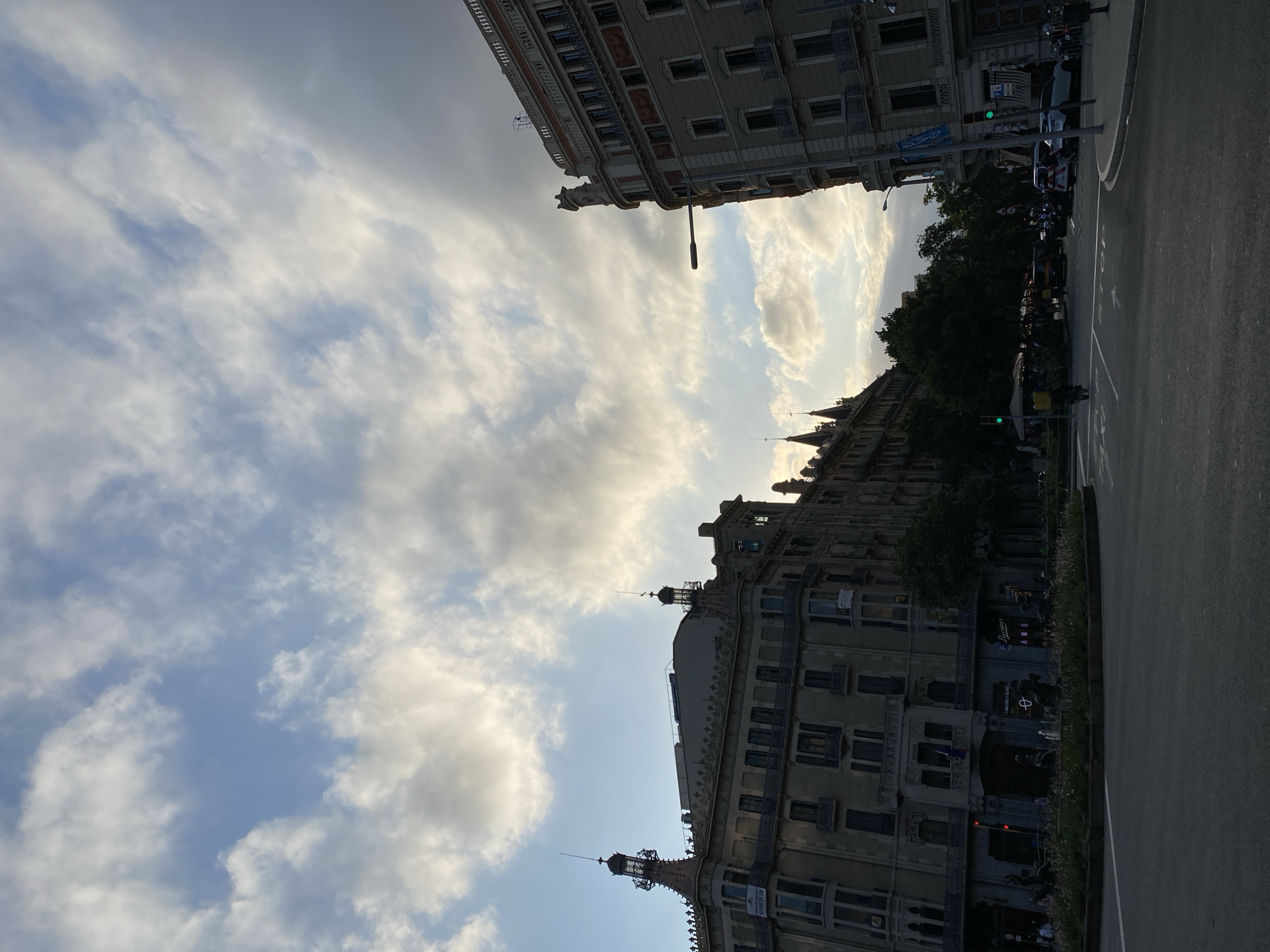 This is a street-level, eye-level photograph taken in Barcelona, Spain, on a cloudy day. The perspective is from the center of a street intersection, looking up towards two imposing buildings that flank the view. The building on the left is a darker, ornate structure with a distinctive mansard roof and decorative spires. The building on the right is lighter in color and has a more classical architectural style.
The sky is filled with a dramatic display of clouds, with patches of blue sky visible. Sunlight is breaking through in some areas, illuminating the edges of the clouds and creating a soft, diffused light. Trees line the street between the buildings, their dark foliage contrasting with the lighter sky.
In the foreground, the asphalt road is visible, with lane markings and a stop sign. Cars and a scooter are present in the scene, suggesting normal city traffic. Pedestrians can be seen on the sidewalks and near outdoor seating areas in front of the buildings. The overall impression is a dynamic urban environment under a dynamic sky.