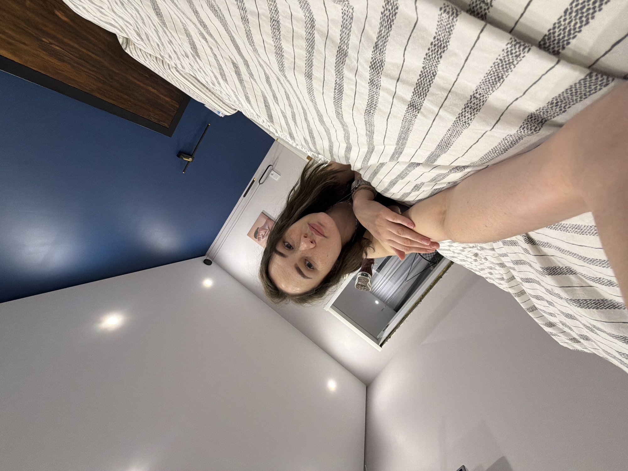 A young woman is depicted lying on a bed, looking directly at the camera from a low angle. She has long, dark hair, fair skin, and noticeable freckles, with a neutral expression. She is partially covered by a white duvet or blanket featuring thin, dark gray striped patterns. Her left arm, adorned with a watch, rests on the bedding, while her right arm is bent with her hand near her chest.
The bedroom features a deep blue accent wall situated behind a dark wooden headboard. The ceiling and other visible walls are painted white. Several illuminated recessed spotlights are present on the white ceiling, casting bright reflections. A small, framed picture showing two indistinct figures hangs on a white wall below the blue accent. A dark, thin fixture, possibly a lamp or decorative item, is mounted on the blue wall. In the background, past the woman's head, an open doorway or alcove with white trim reveals a glimpse into another area, possibly showing a window or fan. The illuminated lights suggest the photo was taken indoors with artificial lighting. The scene is set in Chaikovsky, Russia. No visible text is present.