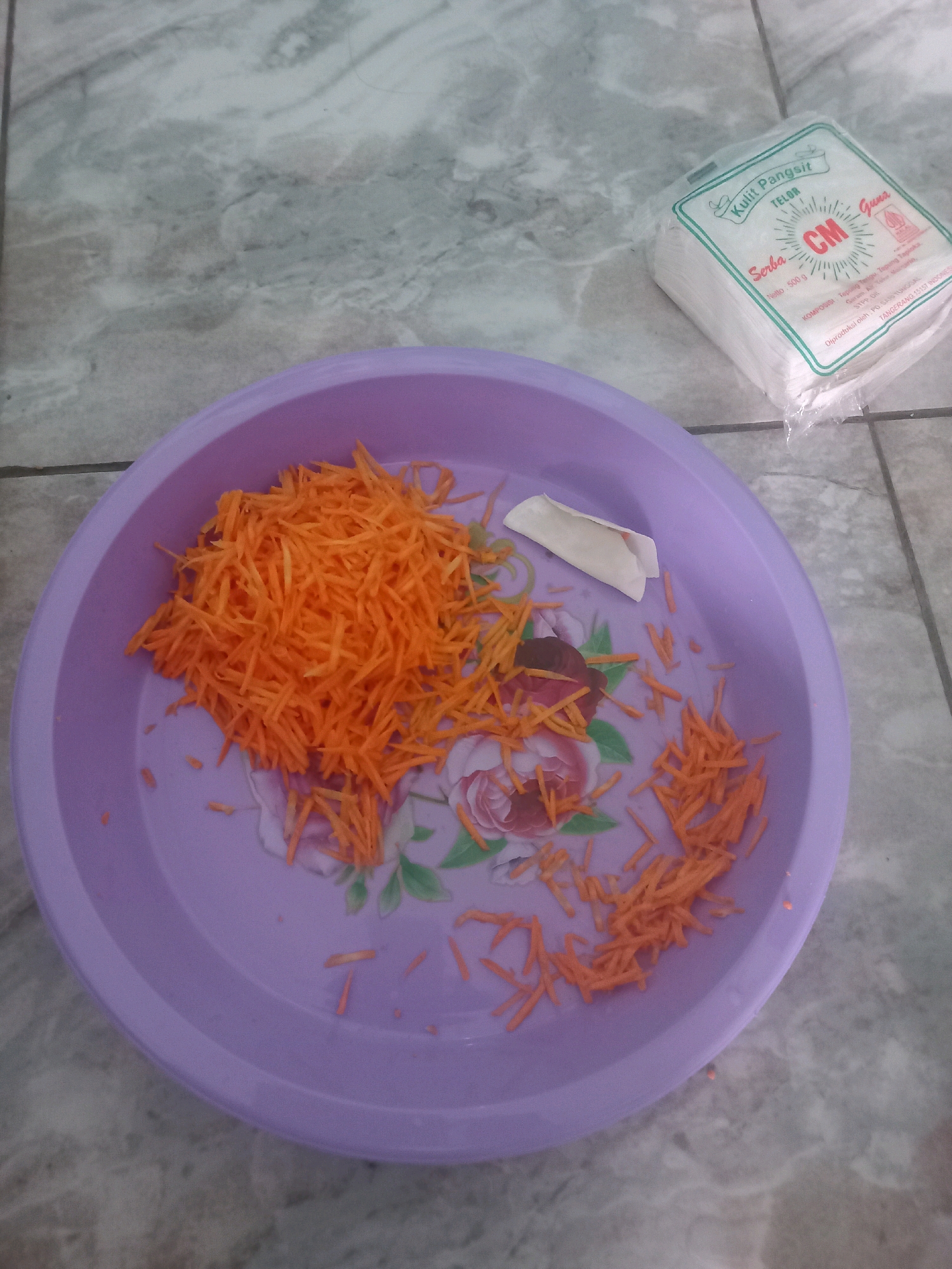 In a tiled room in Depok, Indonesia, a light purple plastic bowl sits on the floor, filled with grated carrots. To the right of the bowl is a white package of spring roll wrappers, labeled "Kulit Pangsit TELOR" and "CM Guna". A few scattered pieces of grated carrot are also visible on the floor around the bowl. The floor is tiled with a gray and white marble pattern.