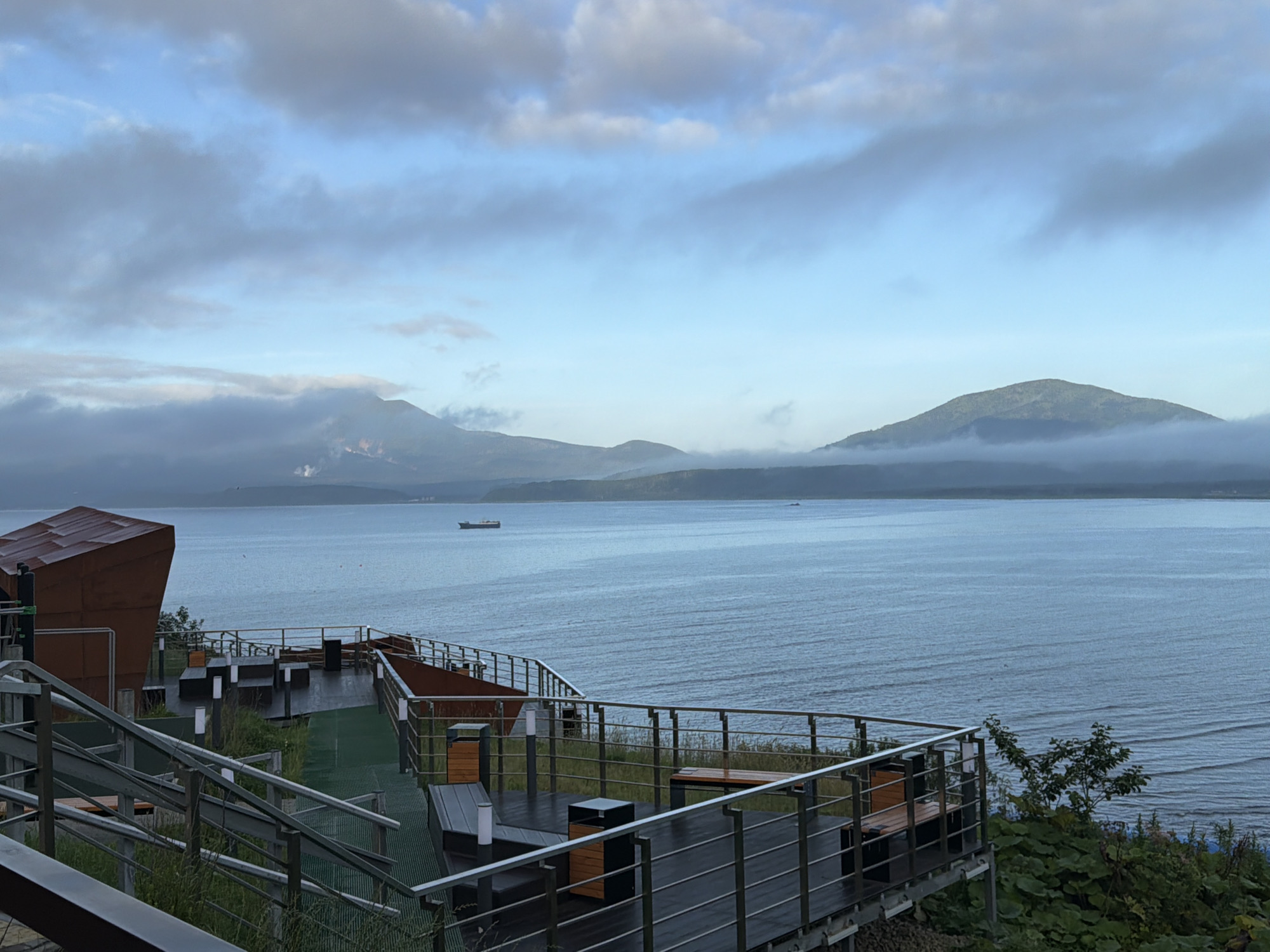 The image shows a view of the Sea of Okhotsk from an elevated outdoor seating area. In the foreground, there is a modern structure with metal railings and wooden benches and tables. The seating area is partly covered with green turf. In the middle ground, the calm sea stretches out, with a cargo ship visible in the distance. In the background, there are rolling hills or mountains, partially obscured by clouds. The sky is a mix of blue and gray clouds, suggesting an overcast day. The location is identified as Yuzhno-Kurilsk, Russia. There are no people visible in the image. The scene appears to be a restaurant or cafe with an outdoor seating area overlooking the sea.