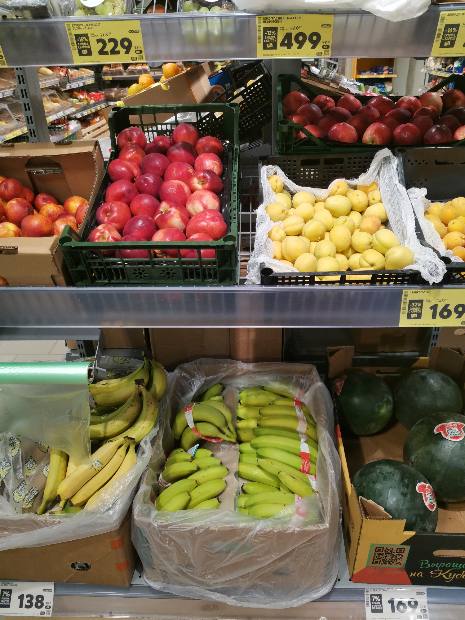 This is a medium shot, eye-level, indoor photograph of a supermarket produce aisle. The focus is on the shelves displaying various fruits, with a price tag visible in the foreground.
On the top shelf, there are crates of red apples and yellow apricots. The apples are in a dark green plastic crate. The apricots are in white plastic bags within a dark green plastic crate.
On the shelf below, two large cardboard boxes contain bananas. The box on the left holds a bunch of yellow bananas. The box on the right contains several bunches of green bananas. Next to the banana boxes are two whole watermelons. The one on the left is partially visible, and the one on the right is fully in view, showing a red sticker with Cyrillic text.
A price tag in the lower right corner displays "169" with "99 P" below it and "КГ" to the right. Another price tag on the left shows "138" with "99 P" below it and "кг" to the right. There are also price tags above the fruits on the shelves, indicating prices and discounts. The environment appears to be well-lit. No people are visible in the image.