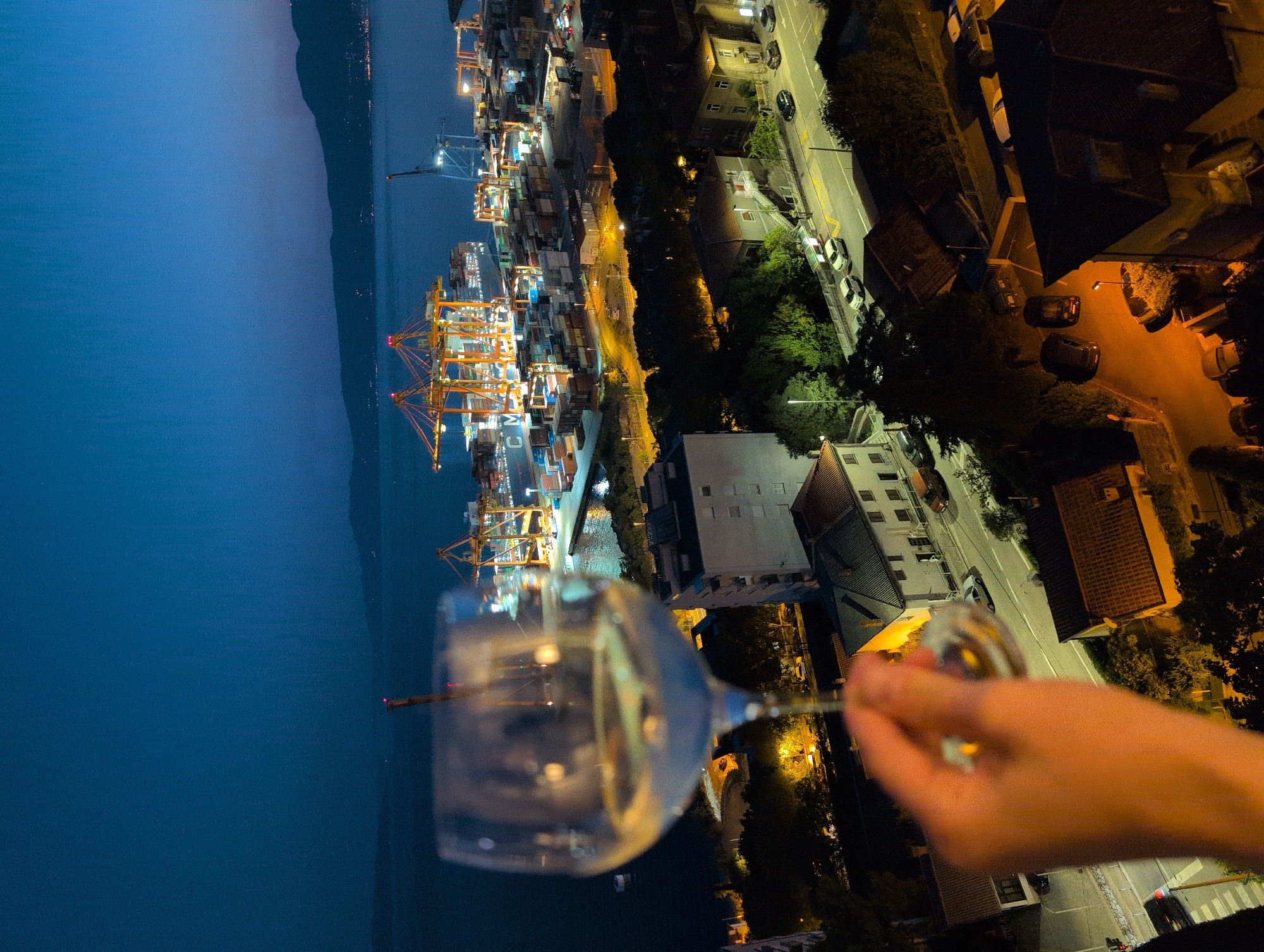 A hand holds a wine glass with white wine in the foreground, obscuring a view of the city of Rijeka, Croatia at night. Beyond the glass, a large industrial port is illuminated with bright lights, featuring numerous cargo containers stacked high and several towering cranes. The port is situated on the water, with a distant shoreline and mountains visible in the background. The foreground shows a residential area with houses and streetlights, indicating an urban setting. The sky is dark blue, suggesting it is nighttime.