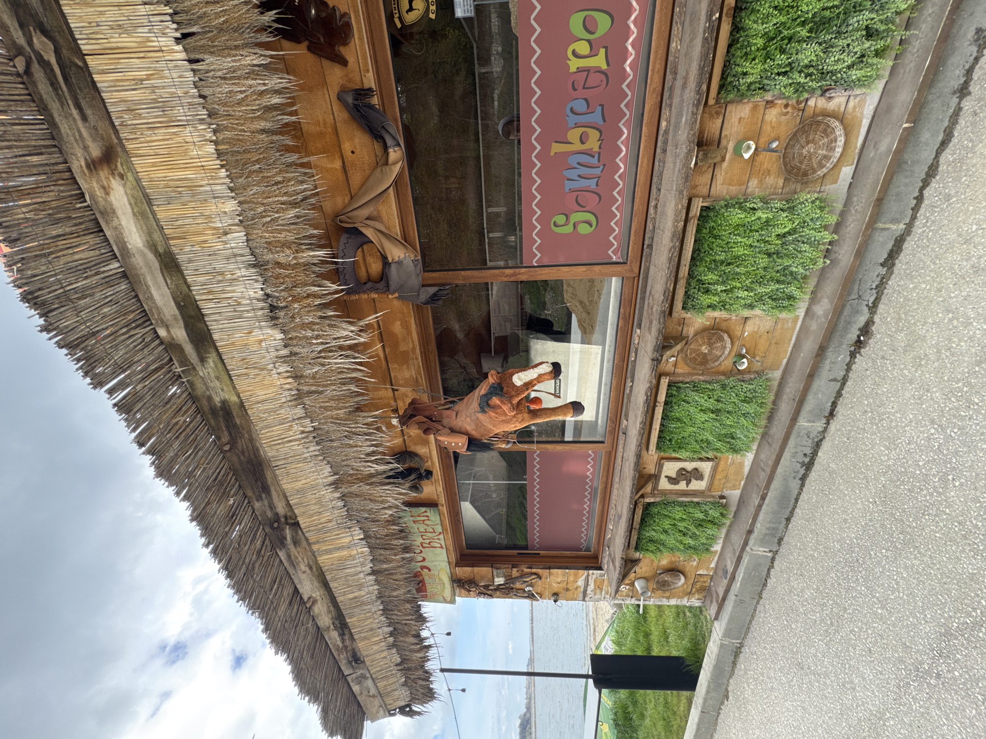 The image shows the exterior of a building with a rustic, possibly themed, design. The building has a thatched roof. The windows have wooden frames and signage, including the word "Sombrero" spelled in bright colors. A stuffed horse with a saddle is mounted on the building facade. Decorative plants are in boxes affixed to the building's wall. The building is located near a body of water. Given the presence of signage in Cyrillic script and the context clues, it's likely located in Varna, Bulgaria. The image includes a concrete sidewalk or road. No people are clearly visible, though there may be a reflection in the window.