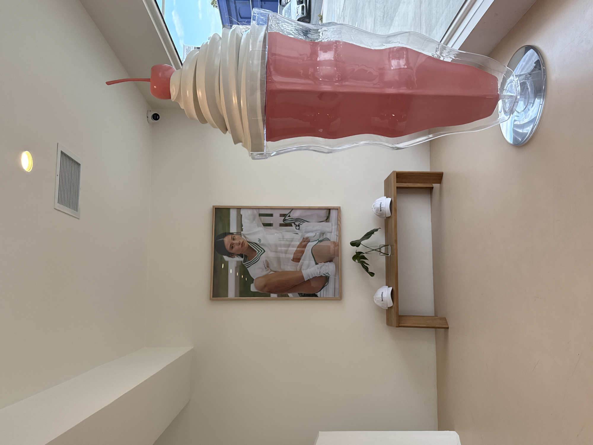 The image captures an interior scene in West Hollywood, California, United States of America. Dominating the upper portion of the frame is an oversized, decorative, clear plastic milkshake glass filled with a pink-colored substance and topped with sculpted white whipped cream and a cherry. The milkshake is positioned near a window, casting a slight shadow on the adjacent wall. Beneath the milkshake, a framed photograph of a woman in athletic attire hangs on the wall. To the right of the photograph is a small, wooden shelf holding a glass vase with a plant and a white hat. The walls are painted in light, neutral tones. Below the photograph and shelf, a white surface is visible, possibly a counter or seating area. A small security camera is mounted on the wall above the photograph and to the left of the decorative milkshake. An air vent is visible on the upper left corner of the frame.
