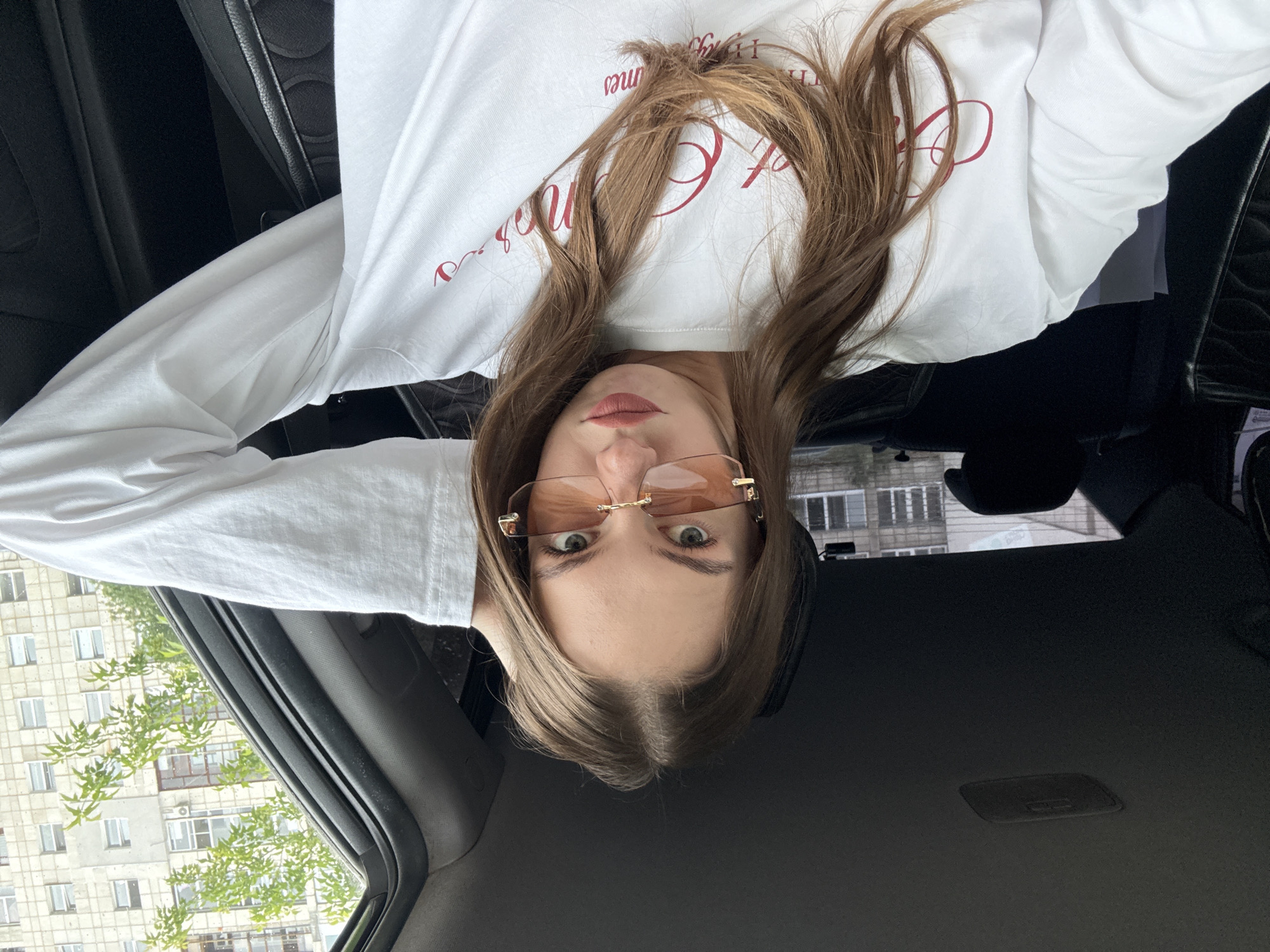 A young woman with long brown hair and light green eyes sits in the driver's seat of a car. She is wearing oversized, hexagonal, rose-tinted sunglasses and a white long-sleeved shirt with red script writing on it. Her right hand is raised and resting on her head. The interior of the car is dark, with black leather seats visible. Through the open car window, a light-colored, multi-story apartment building with many windows and air conditioning units can be seen, along with green trees. The sky is overcast. The setting is likely a residential area in the city of Perm, Russia.