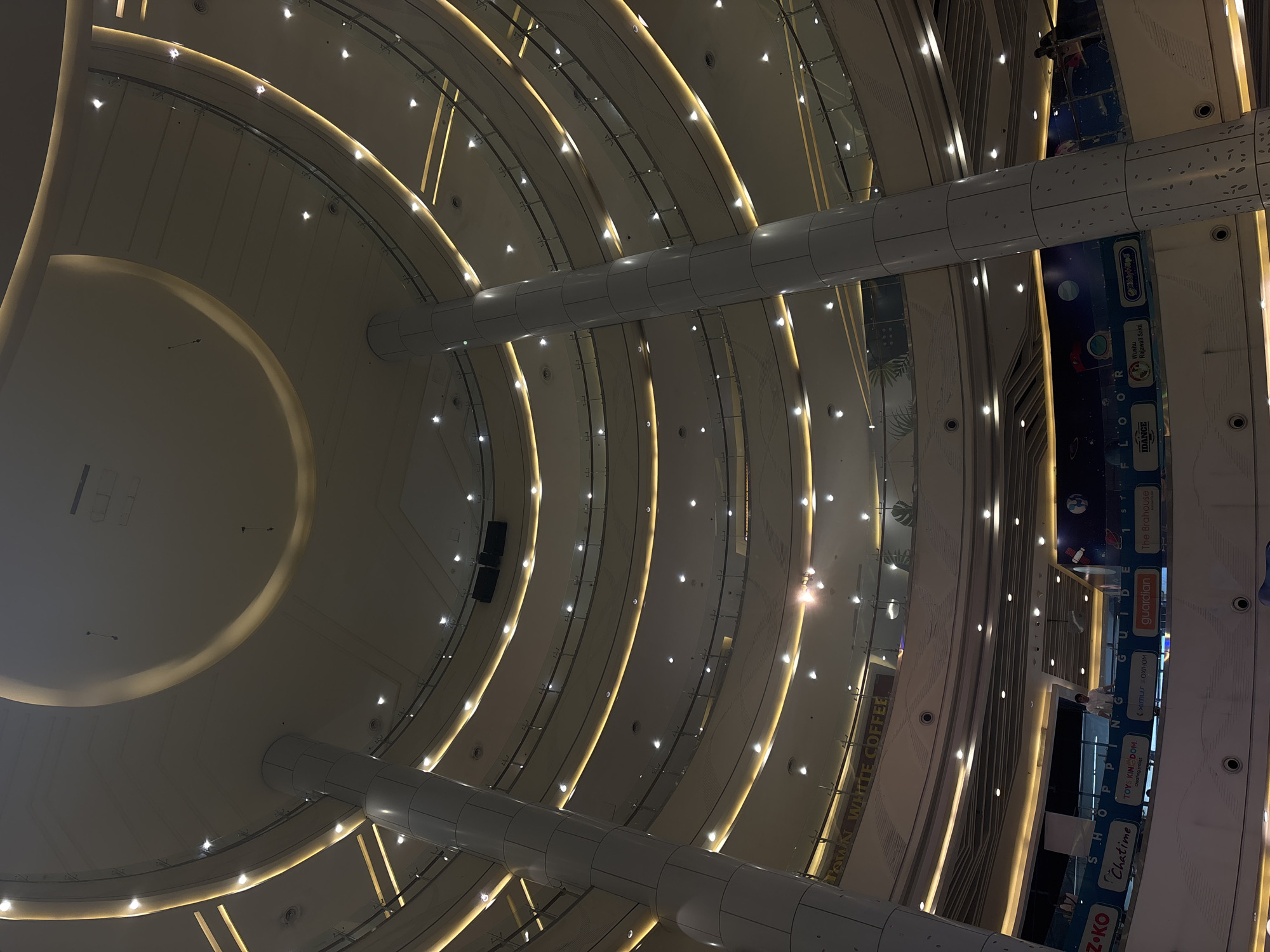 The image displays the interior of Mall Artha Gading, a multi-story shopping mall located in North Jakarta, Indonesia, viewed from a lower level looking upwards into its vast circular atrium. Numerous concentric floors ascend, each distinguished by light-colored ceilings featuring recessed spotlights and continuous warm yellow LED strip lighting along the edges. Glass railings with metallic handrails define the perimeter of each level. Two prominent, light-grey, segmented cylindrical pillars extend vertically through the center of the atrium. Branding visible on a lower floor banner includes "Chatime," "TOY KINGDOM," "SHIMIZU OXIHOM," "guardian," and "The Brahouse," alongside "Wushu Rajawali Sakti." "OLD TOWN WHITE COFFEE" is also discernible on an upper floor. A single male individual in a light shirt stands leaning against a railing on a middle floor. The environment is modern, spacious, and illuminated by extensive artificial lighting.