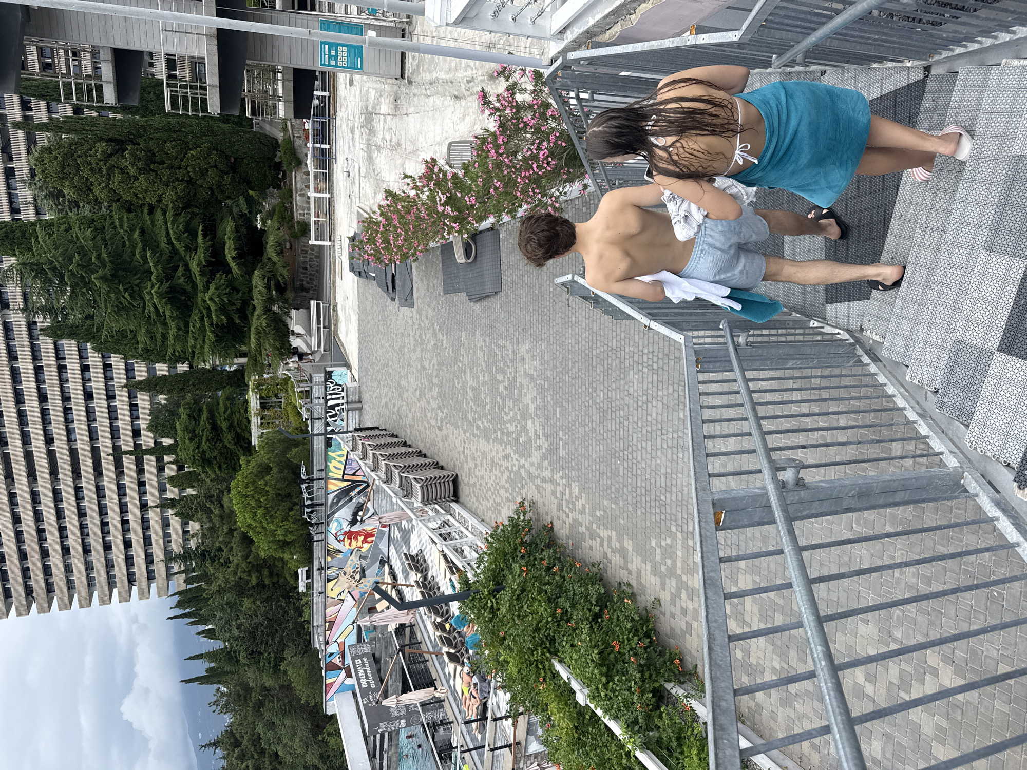 In the city of Yalta, Ukraine, a young shirtless man and a woman with wet hair wearing a white bikini top and a turquoise towel are descending a metal staircase. They are both carrying towels. The staircase leads down to a paved area next to a swimming pool area with lounge chairs and umbrellas. In the background, a large, multi-story building with numerous windows is visible, surrounded by lush green trees. The sky is overcast, and the overall atmosphere suggests a resort or hotel setting.