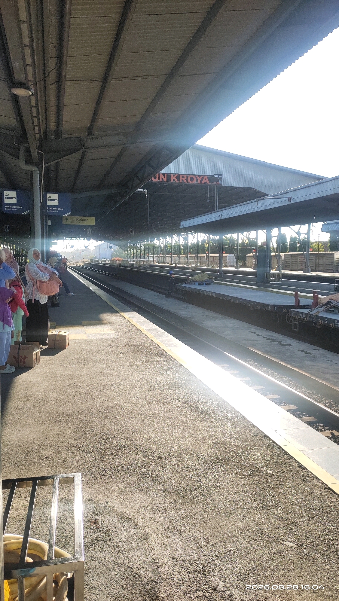 The image captures a sun-drenched scene at a train station platform in Kroya, Indonesia. The perspective is from ground level, looking down a set of train tracks under a covered walkway. The walkway's corrugated metal roof casts shadows, with bright sunlight streaming through the gaps and from the right side of the frame, creating a lens flare effect.
On the platform to the left, a group of people are visible. A woman wearing a light-colored headscarf and a patterned top is holding a baby or a large pink bundle. She is standing near several cardboard boxes. Other individuals in casual attire are also present, some appearing to be waiting or socializing.
The train tracks stretch into the distance, with another platform visible on the right side, also partially covered. Several freight cars are parked on the right-hand track. Signage is visible on the pillars supporting the walkway, including one that reads "Area Merokok" (Smoking Area) and another with an arrow pointing towards "Keluar" (Exit). A large red sign with white lettering on the structure in the background appears to read "UN KROYA".
The ground of the platform is paved with textured material, and a yellow line marks the edge of the tracks. The overall atmosphere suggests a typical day at a provincial train station. The timestamp in the lower right corner indicates "2026.08.28 16:04".