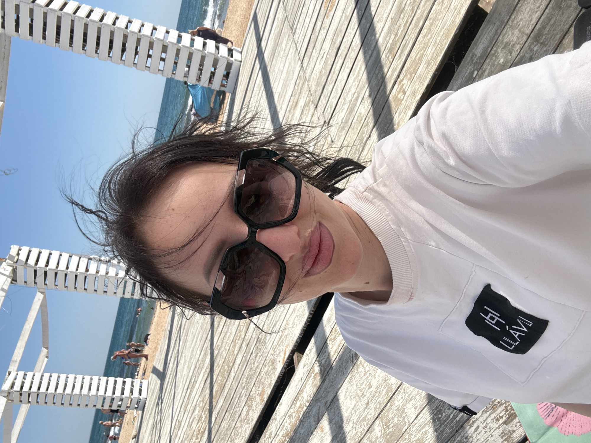 A woman with dark hair and sunglasses takes a selfie on a wooden pier. She is wearing a white t-shirt with a black logo on the left chest. Behind her, the sea and a sandy beach are visible, with several people in the water and on the shore. The sky is blue and clear.