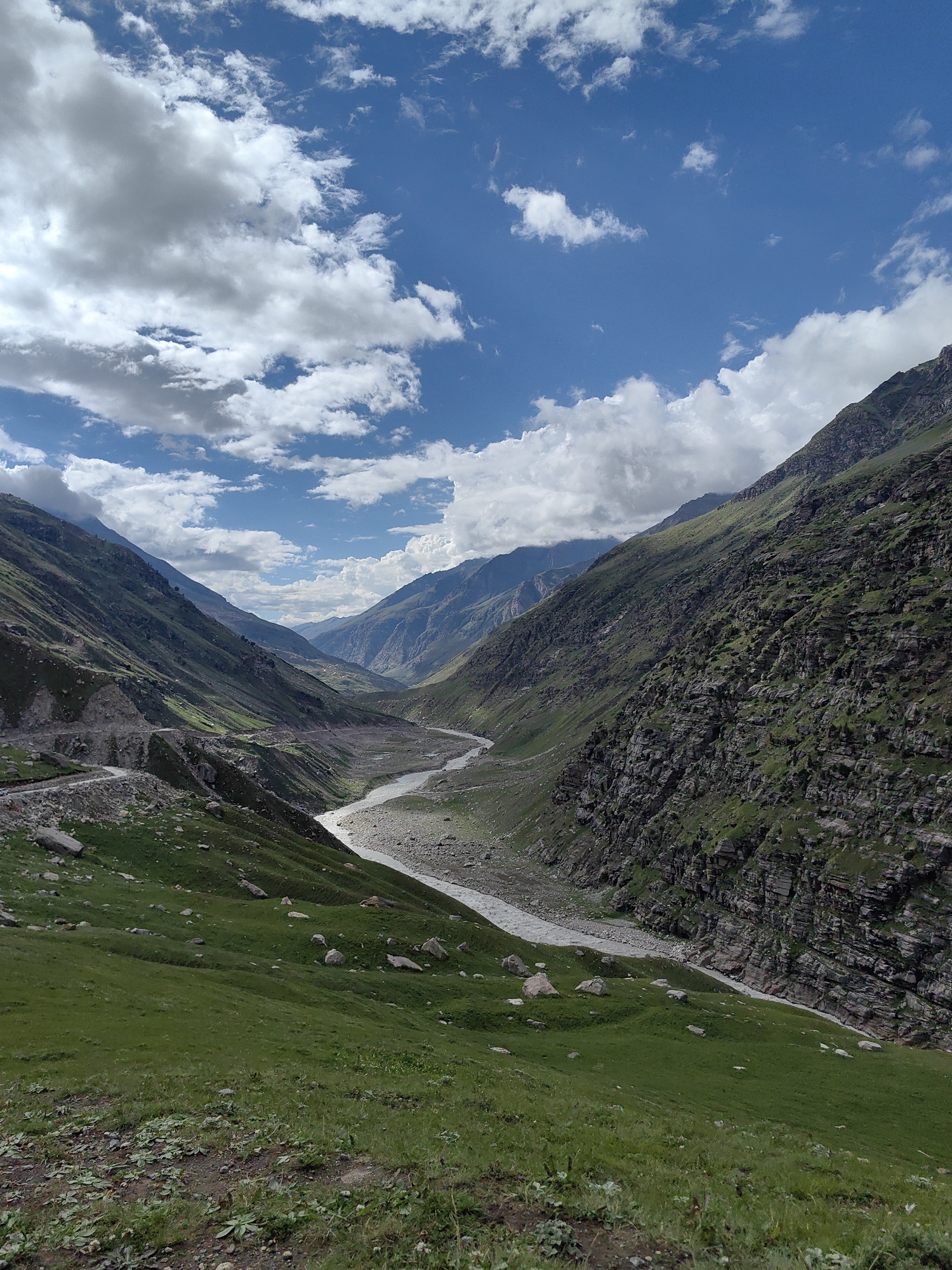 A sweeping, high-angle view of a verdant valley in the Himalayas, near the town of Bashisht, India, under a partially cloudy blue sky. A winding, rocky river carves its way through the valley floor. The slopes of the surrounding mountains are covered in lush green grass and interspersed with rocky outcrops and sheer cliff faces. A narrow, unpaved road is visible winding along the left side of the valley. The scene is one of rugged natural beauty, with no people or man-made structures other than the road visible.