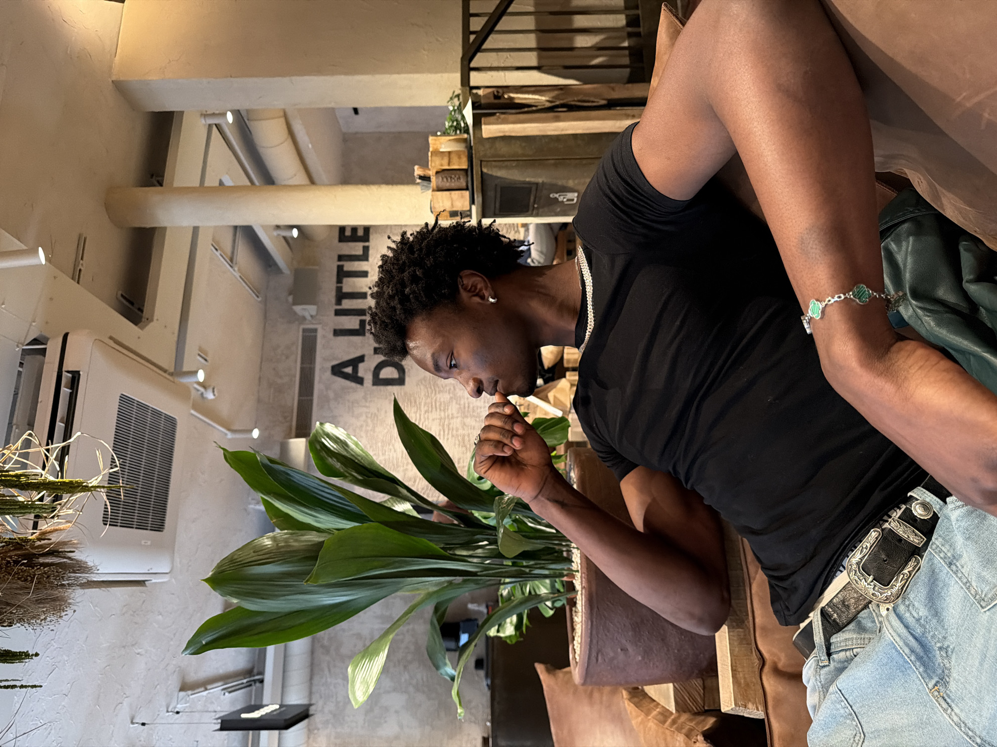 The image shows a person seated in an indoor location. The individual, a dark-skinned person with short curly hair, is wearing a black T-shirt, a silver chain necklace, blue denim jeans with a decorative black leather belt, and a silver clover-shaped bracelet on their wrist. The person is seated near a large potted plant with long green leaves. In the background, there is a wall with partial lettering visible, possibly part of a sign or logo, that includes the words "A LITTLE". Above the plant and sign, there is a commercial air conditioning unit installed on the wall. The overall environment appears to be a cafe or a restaurant. It is not possible to identify the precise location in the city of London, United Kingdom based solely on the visual information.