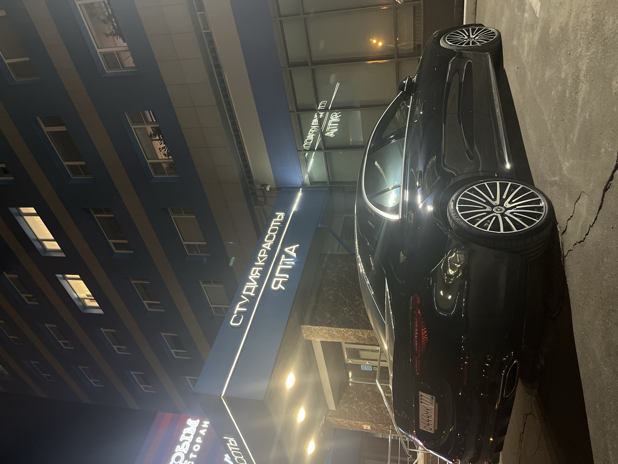 The image shows a black Mercedes-Benz parked on a city street at night in Moscow, Russia. The car is positioned in front of a building that houses businesses, including a beauty salon called "Студия Красоты Ялта" (Studio of Beauty Yalta) and a restaurant called "Новый Ресторан" (New Restaurant). The building is modern, with a facade featuring rows of windows and horizontal stripes. The signage for the businesses is illuminated, creating a bright glow in the immediate vicinity. The street appears to be paved and is slightly cracked or uneven in places. The overall atmosphere is urban and illuminated by artificial lighting. The license plate of the car is partially visible and appears to read "O444MK 777 RUS".