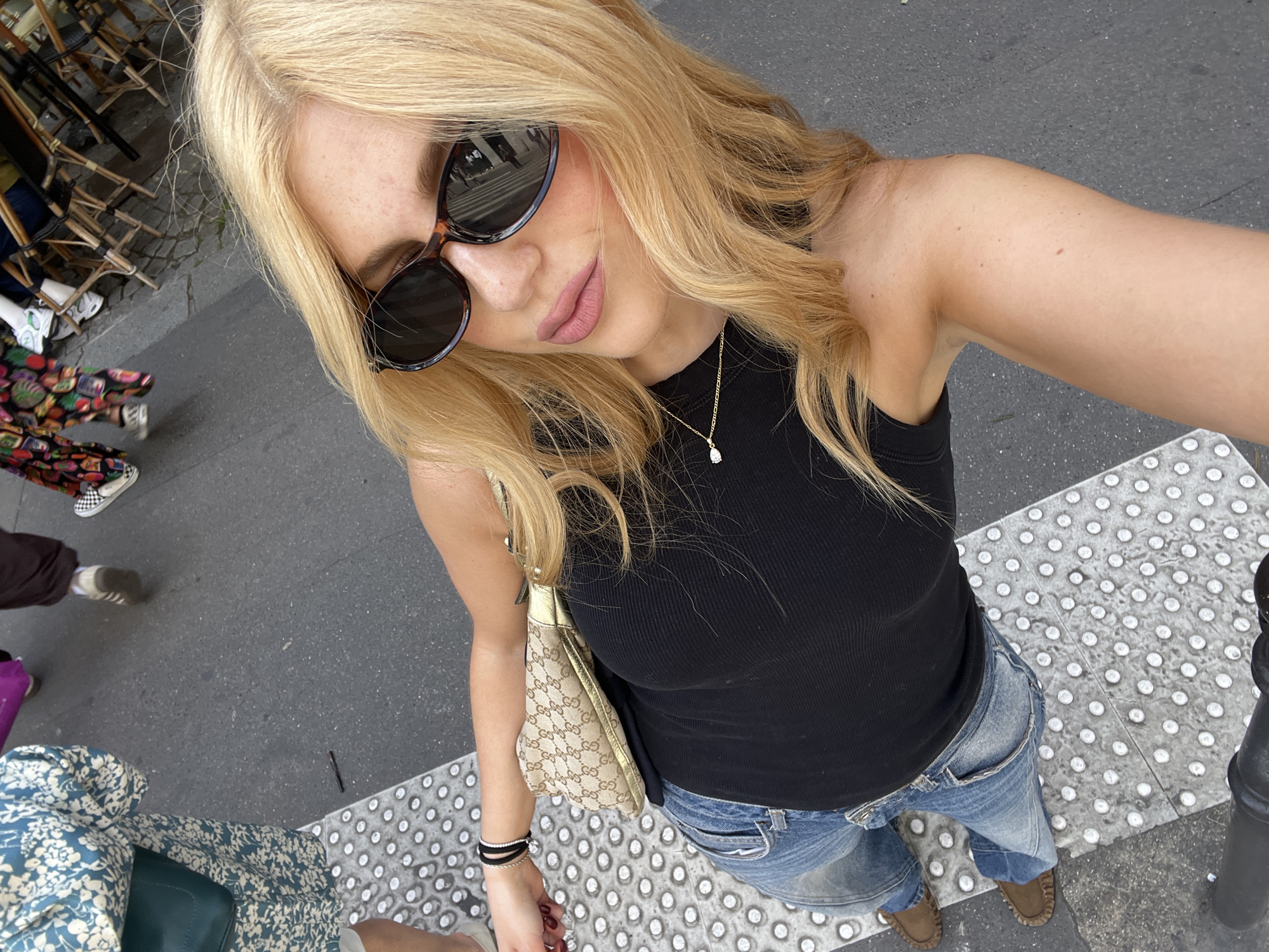 A woman takes a selfie on a Paris, France, street. She is wearing sunglasses, a black tank top, blue jeans, and brown shoes. She has blonde hair and is wearing a gold necklace. She is carrying a gold purse. There are other people walking by in the background. The location is recognizable by the cobblestone street and the typical Parisian chairs.