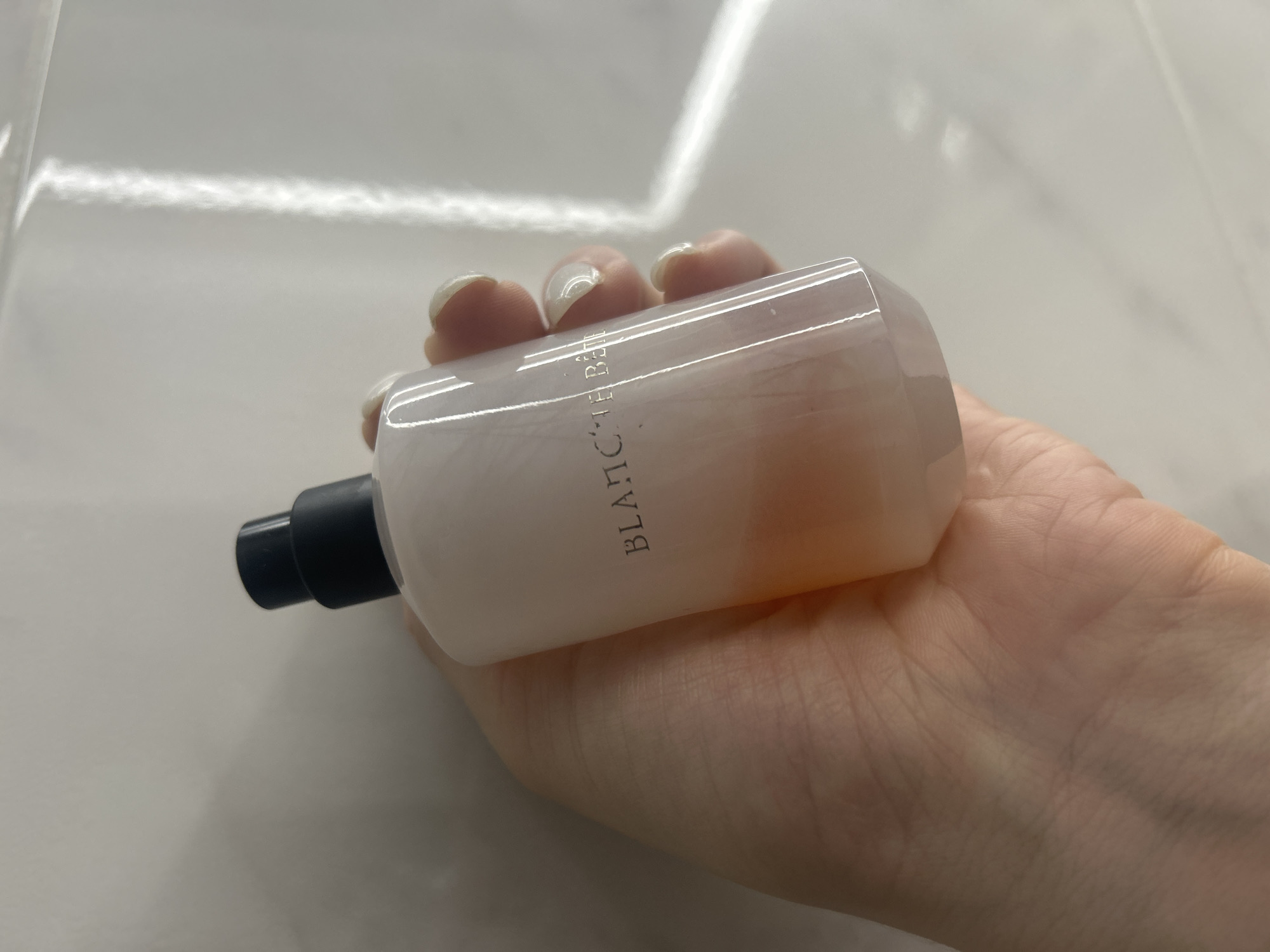 A hand with light-colored, manicured fingernails holds a white, cylindrical bottle with a black spray nozzle. The bottle has a pinkish hue towards the bottom. The text "BLANC le Bain" is embossed in a dark, stylized font on the side of the bottle. The background is a light-colored, textured surface, possibly a tiled wall, with a strong vertical highlight from a light source. The image does not provide any information about the location in the city of Нижние Муллы, Russia.