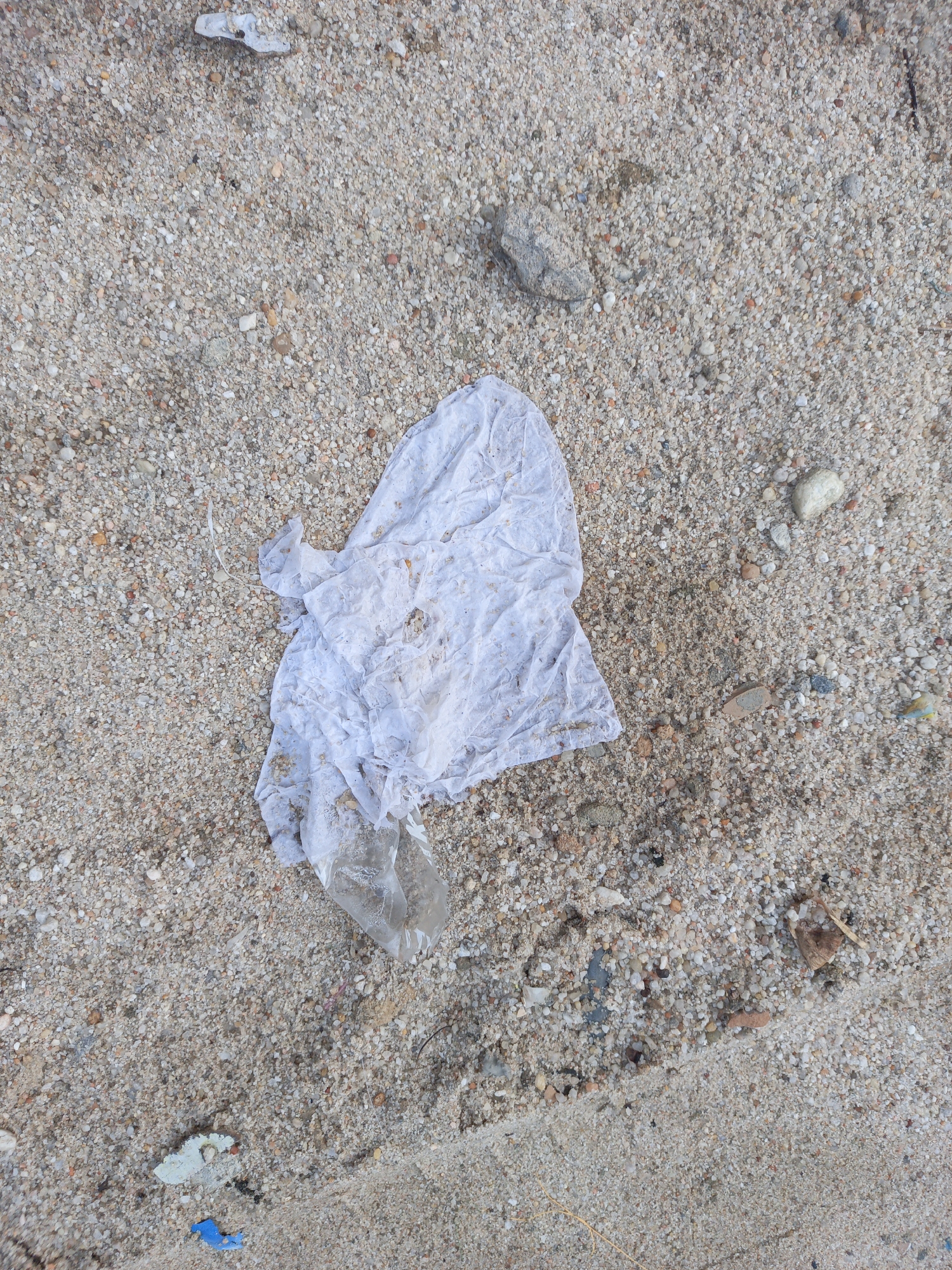 The image shows a close-up of sand with visible grains and small pebbles. A soiled, white cloth is prominently placed in the center. A fragment of transparent plastic is attached to the cloth. Other small pieces of debris are scattered across the sandy surface. The location is Unknown, Unknown. The scene depicts environmental pollution through discarded waste on a beach. No people or interaction are visible.