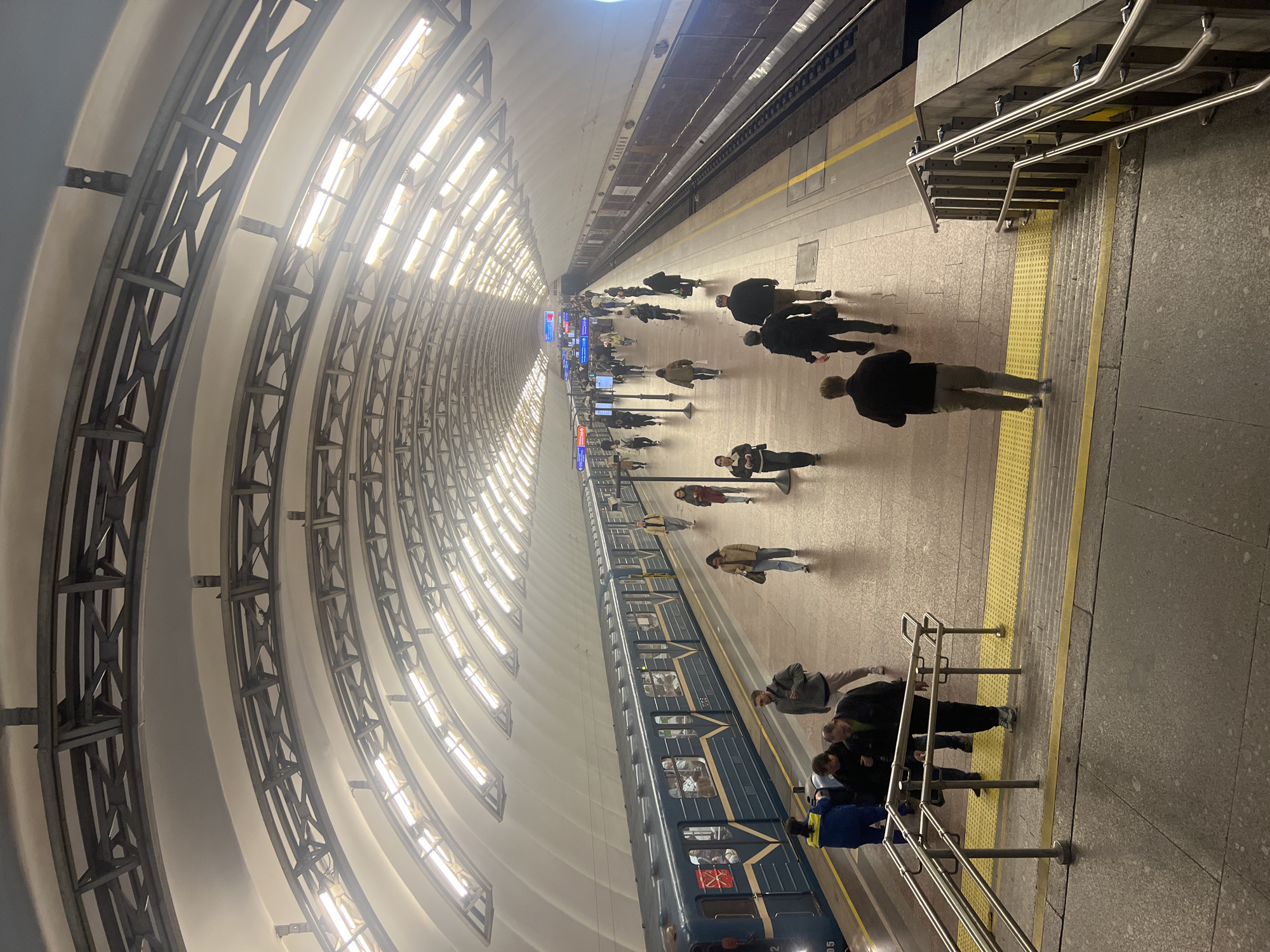 This image depicts a subway station in Saint Petersburg, Russia. The station is characterized by its arched ceiling supported by metal structures and illuminated by rows of lights. A blue subway train is at the platform, with people entering and exiting. Other people are walking along the platform, which has a yellow safety line near the edge. Stairs with metal handrails lead to a higher level. The overall atmosphere suggests a busy, functional public transportation hub.