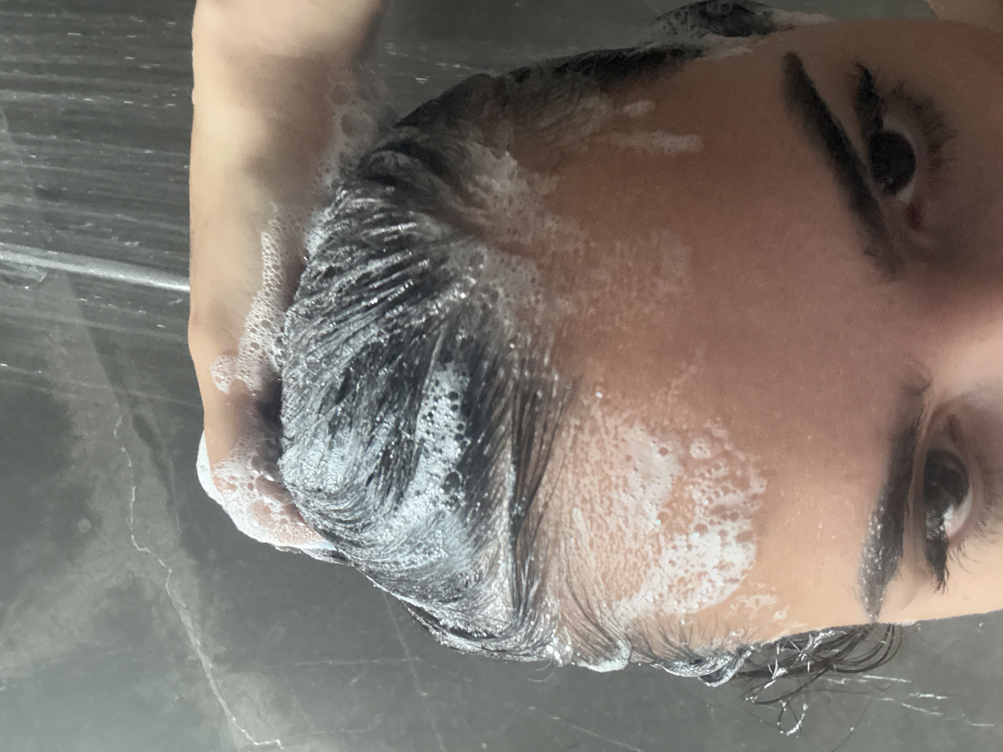 A person is lying on their back in what appears to be a shower or bathroom setting. Their dark hair is wet and covered in soap suds, as is part of their face. The eyes are open and looking toward the camera. The environment is a dark grey surface, possibly tile or marble, which could be the shower floor or bathroom countertop. It's unclear if this image depicts San Bernardino Tlaxcalancingo, Mexico.